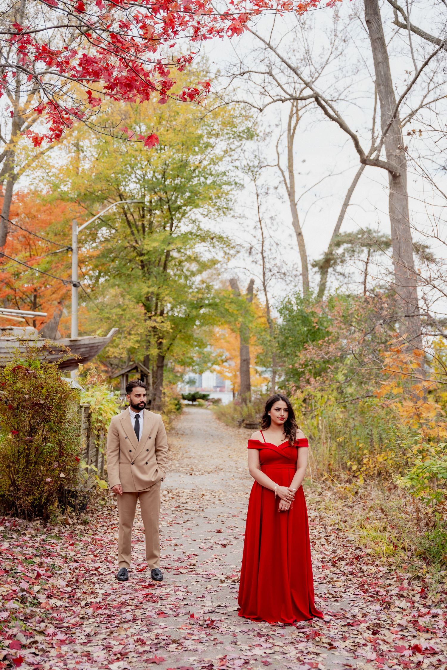 A couple poses in a park path surrounded by vibrant red and orange autumn foliage and fallen leaves.