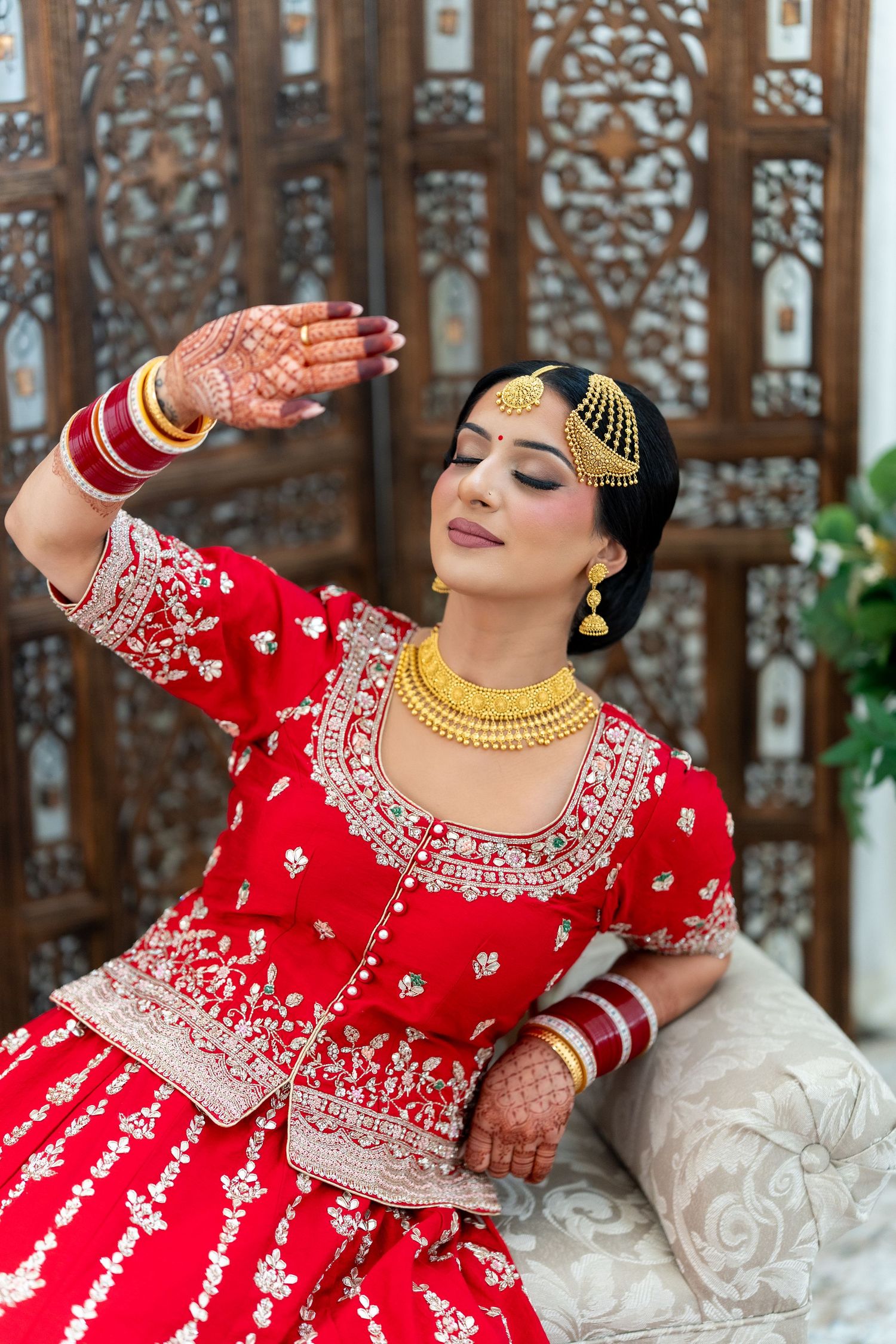Bride in red and gold attire strikes graceful poses on white sofa with decorative wooden panels behind.