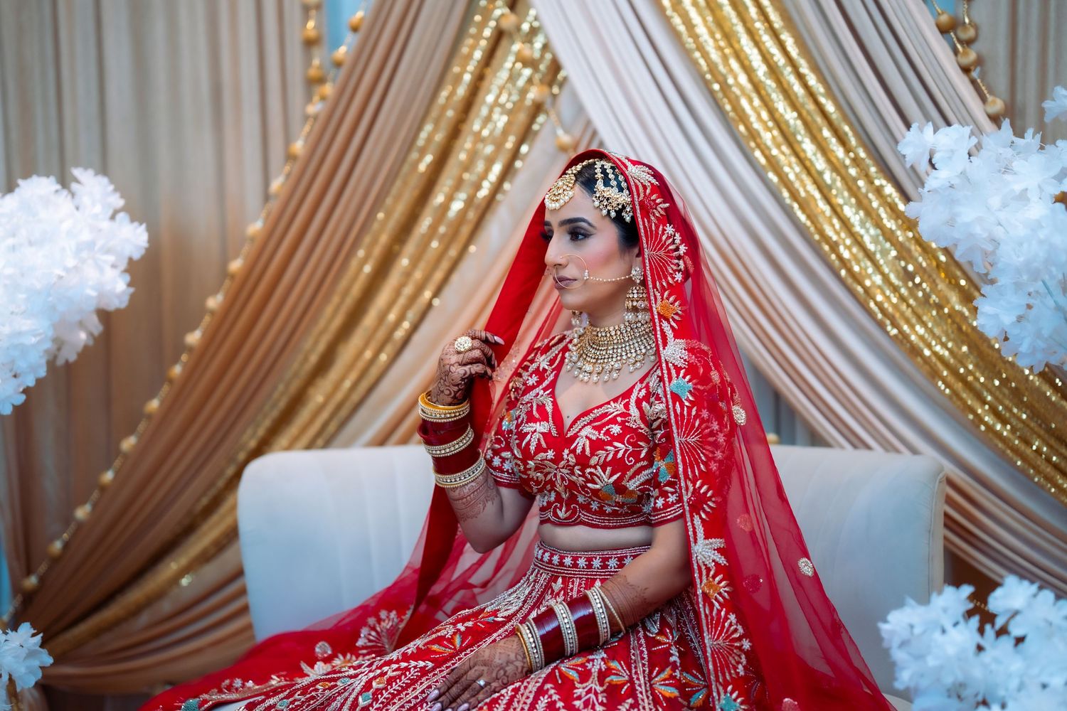 A bride in a traditional red and gold Indian lehenga sits elegantly at her wedding ceremony surrounded by decorative drapery.
