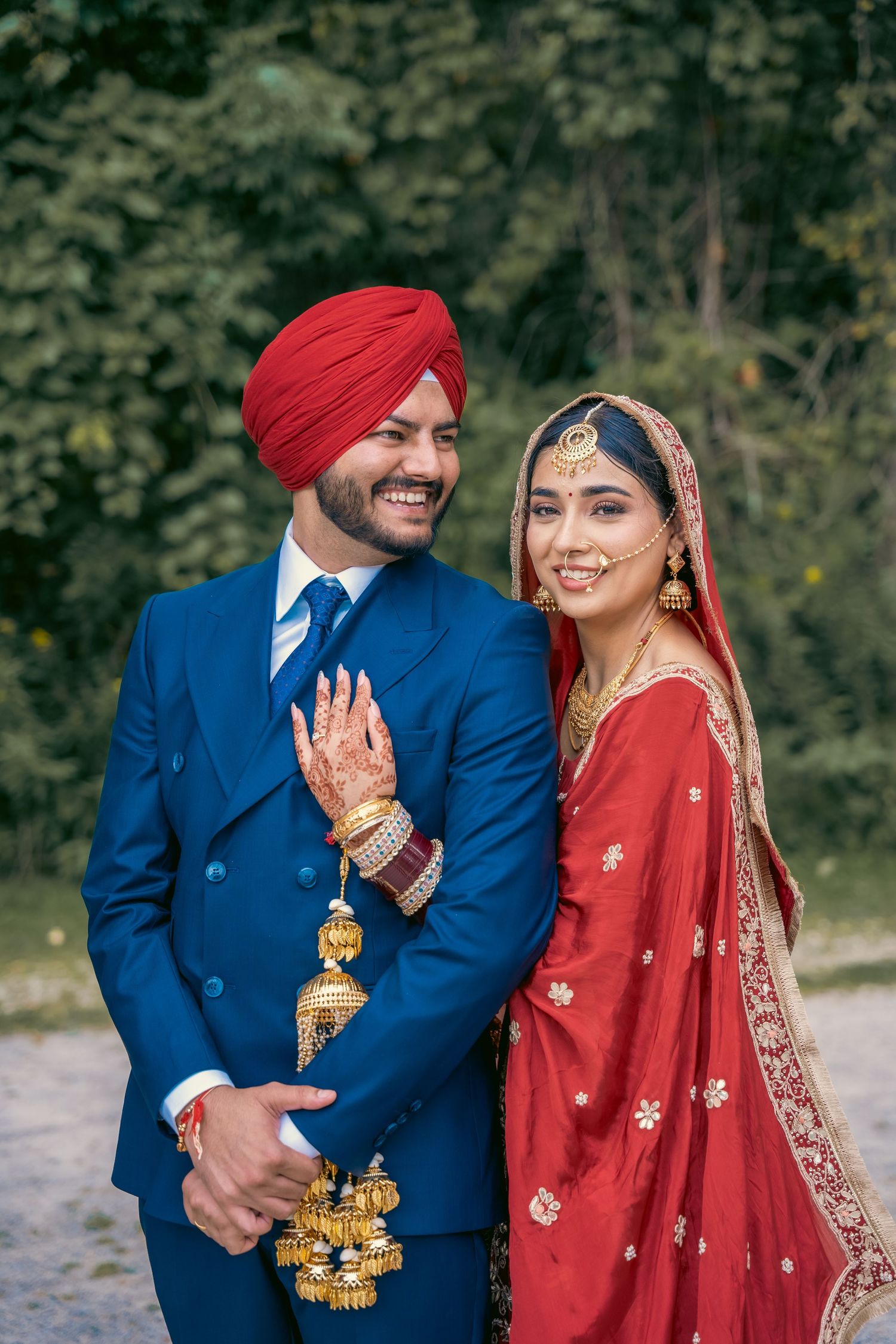 A couple in traditional Indian wedding attire, with the groom in blue and bride in red, pose for romantic outdoor photos.
