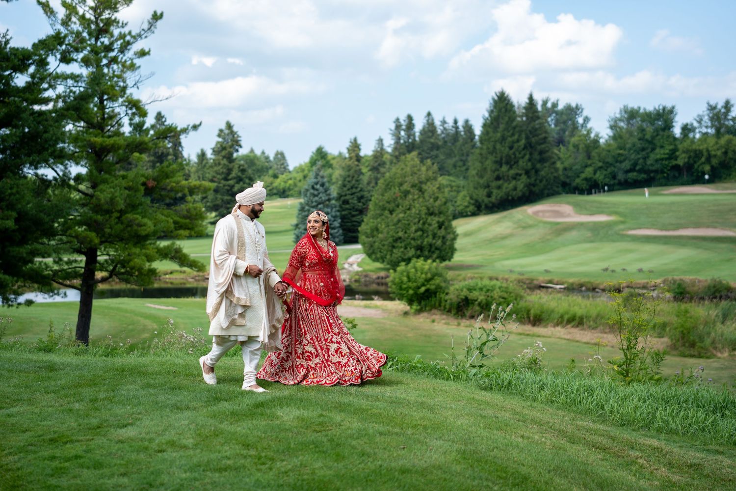 Two people walk together across a scenic golf course landscape with rolling green hills and trees in the background.