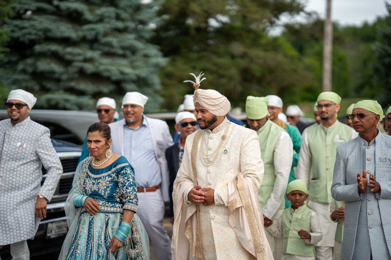 Wedding guests in traditional South Asian attire walking outdoors among evergreen trees.