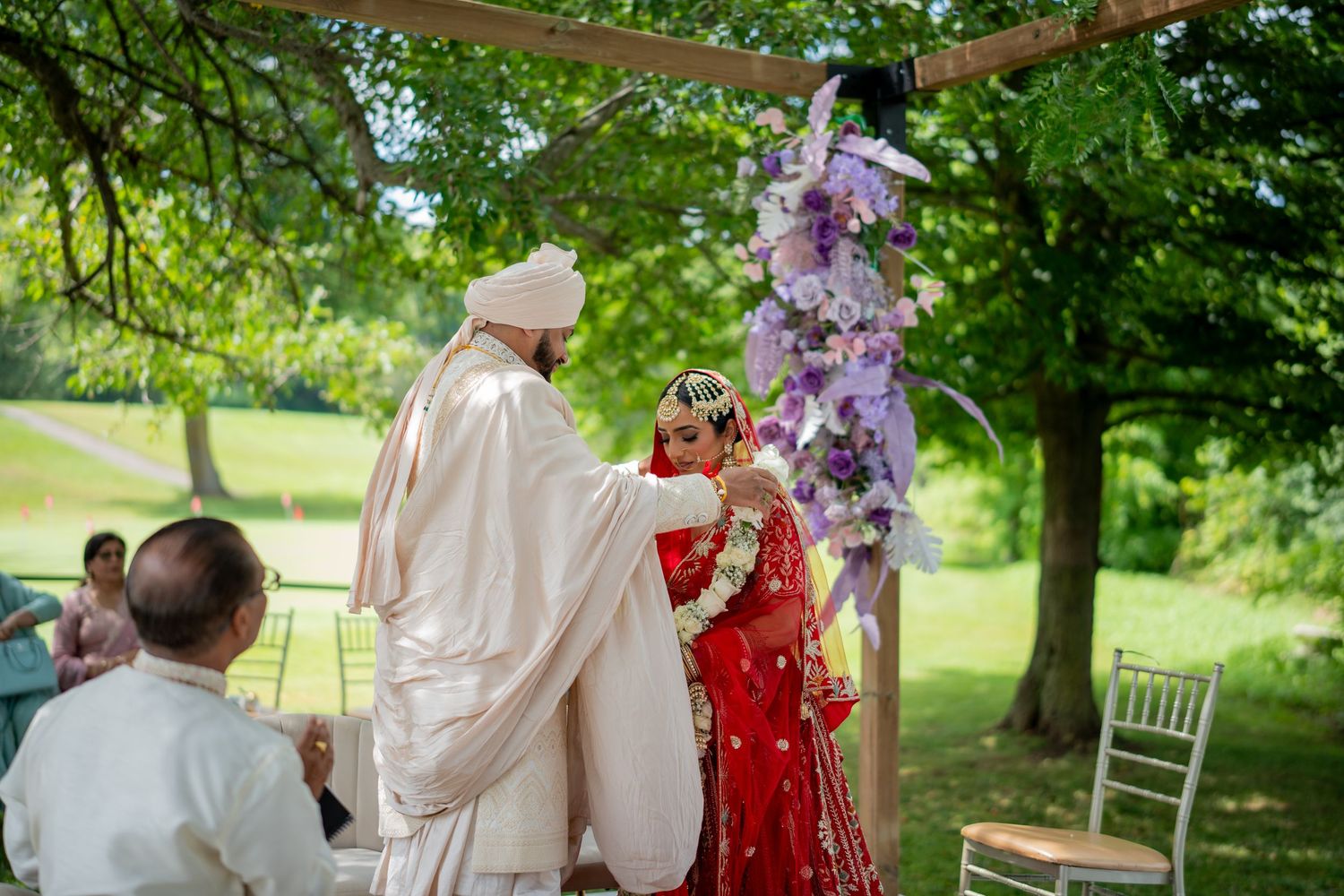 Outdoor wedding ceremony in a lush garden setting with guests gathered under a decorated canopy.
