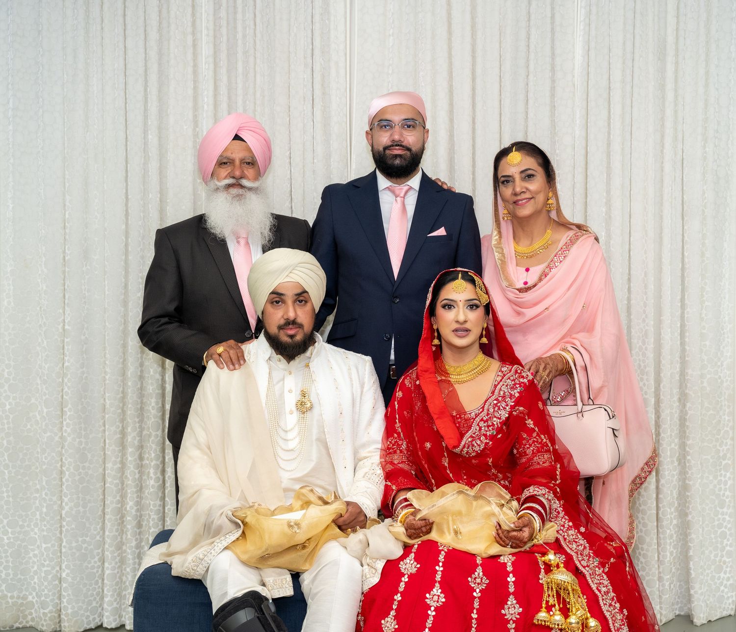 A traditional Sikh wedding party posed together in formal attire against a white backdrop.