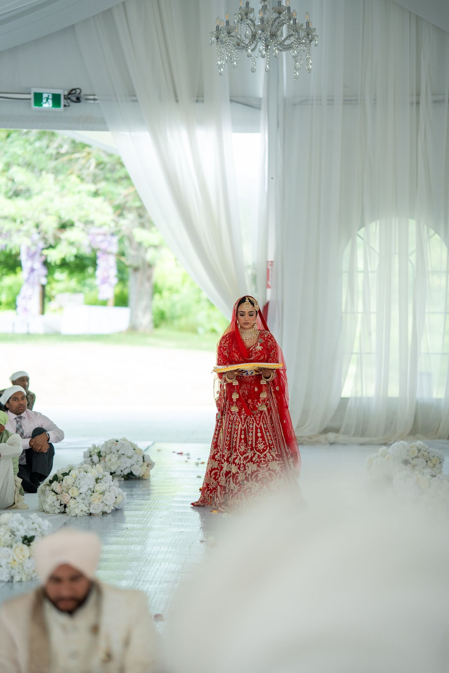 A bride in a stunning red lehenga stands in an elegant white-draped wedding venue with crystal chandelier.