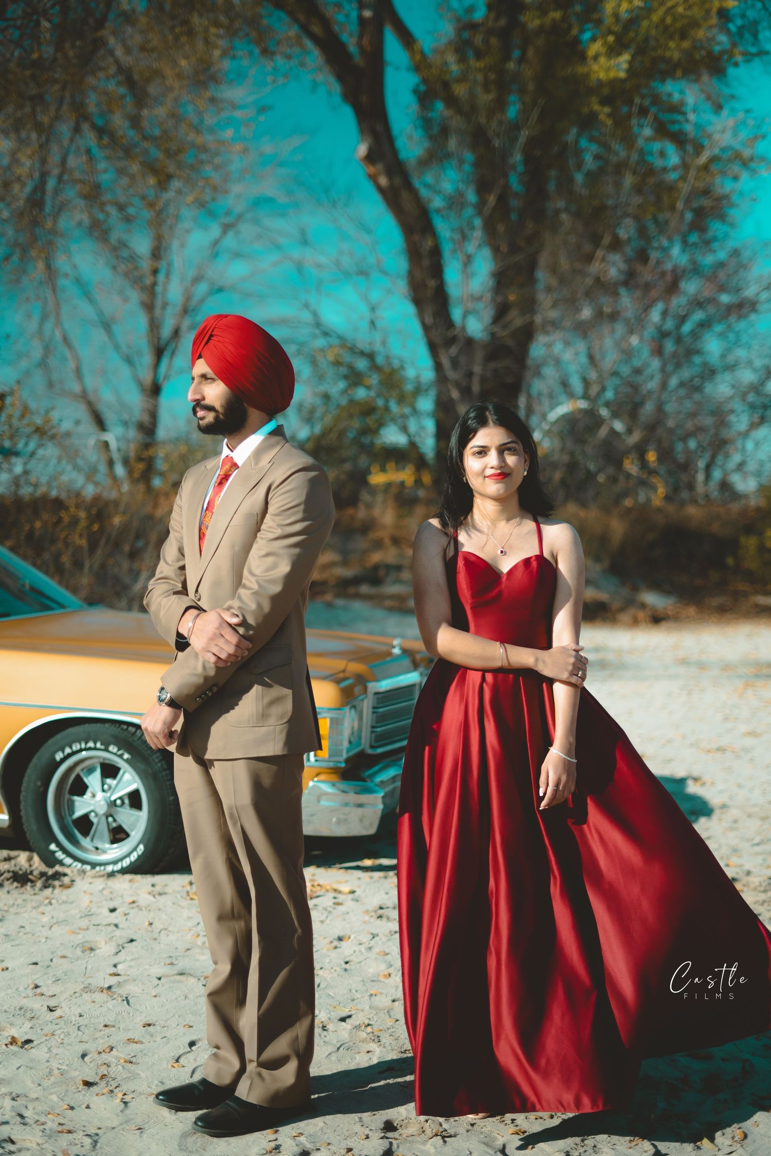 A couple in formal attire poses outdoors with a red dress and tan suit against a dreamy turquoise background.