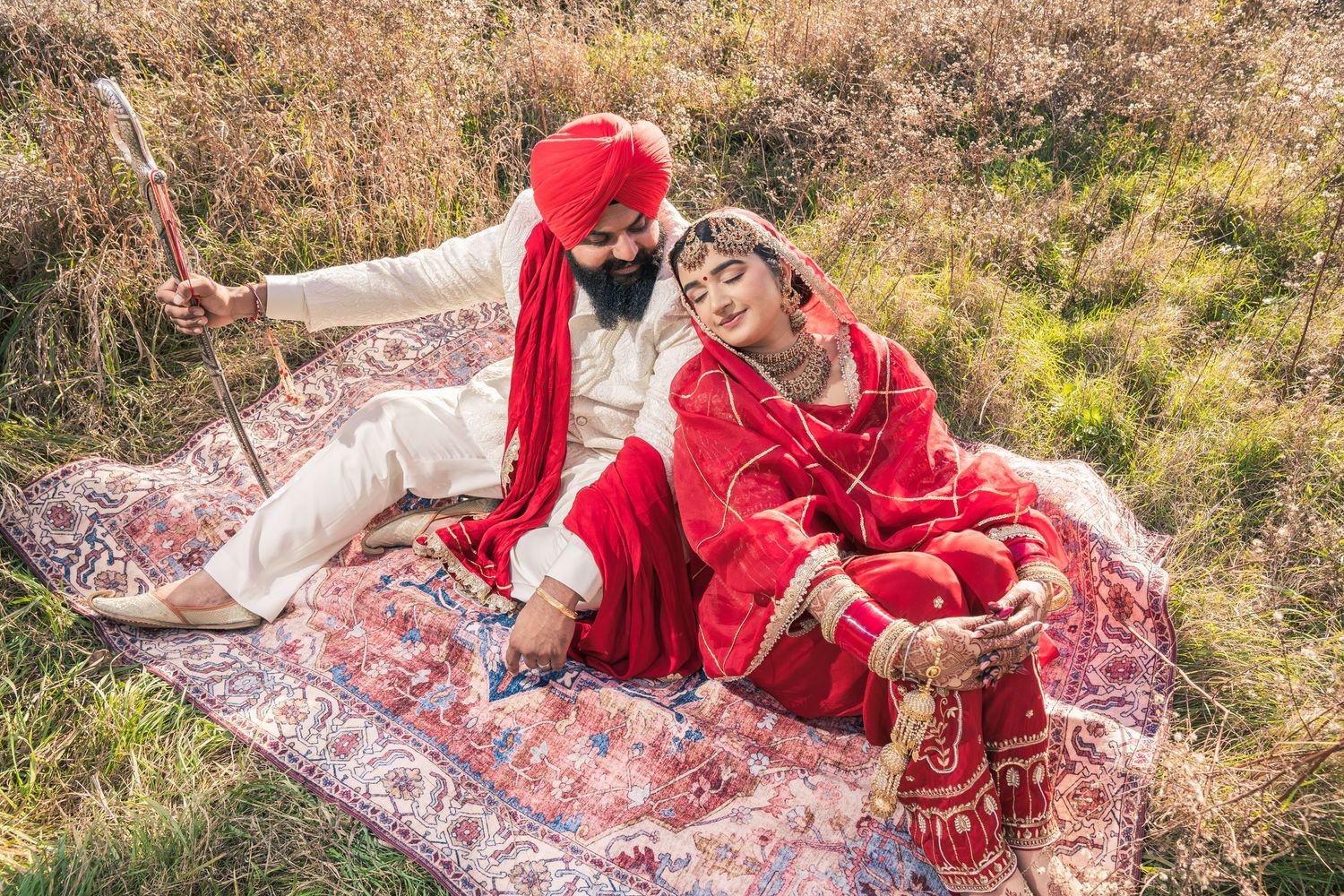 Wedding photoshoot on a decorative rug outdoors with traditional red Indian attire.