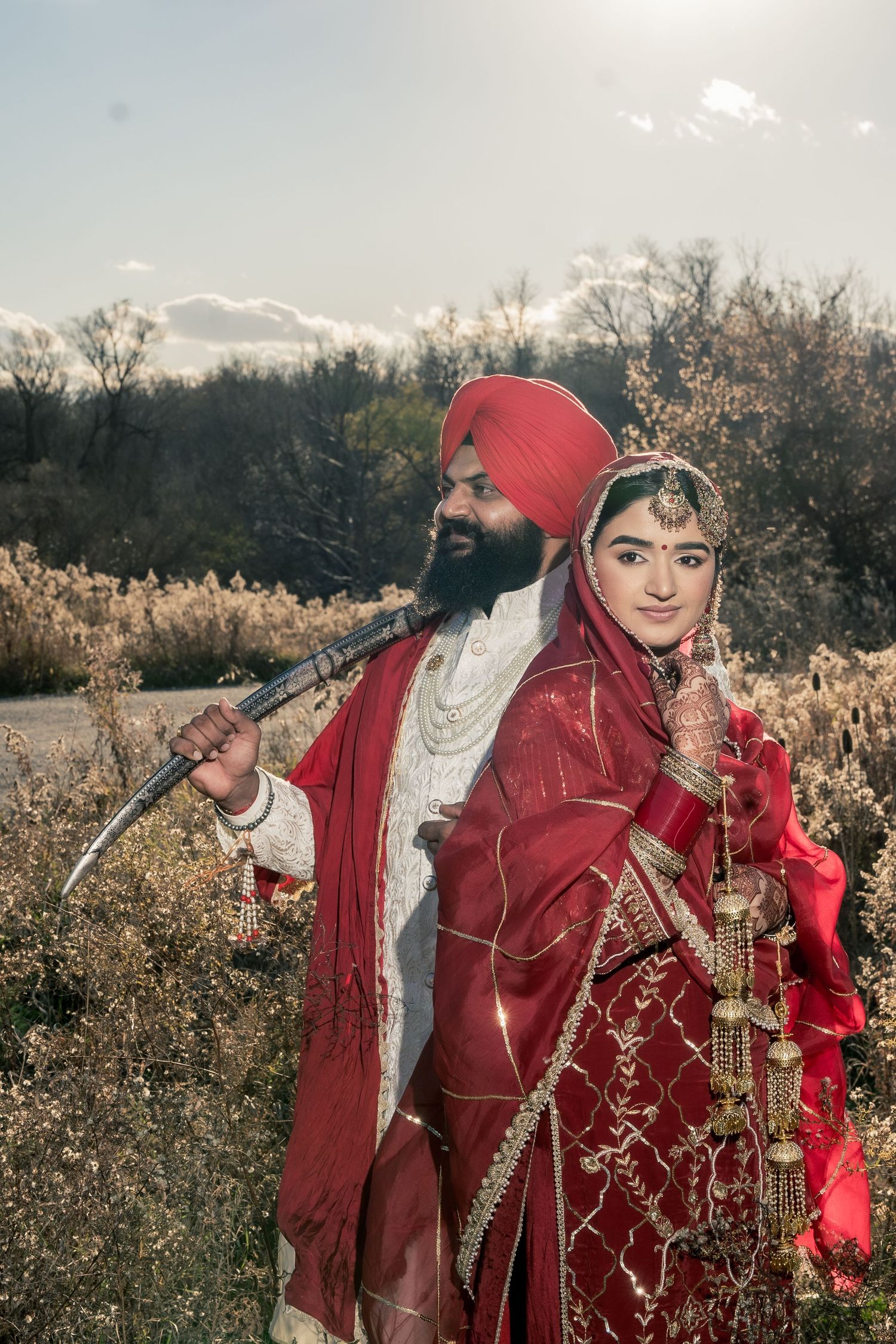 Couple in red traditional Indian wedding attire poses in a field during golden hour.