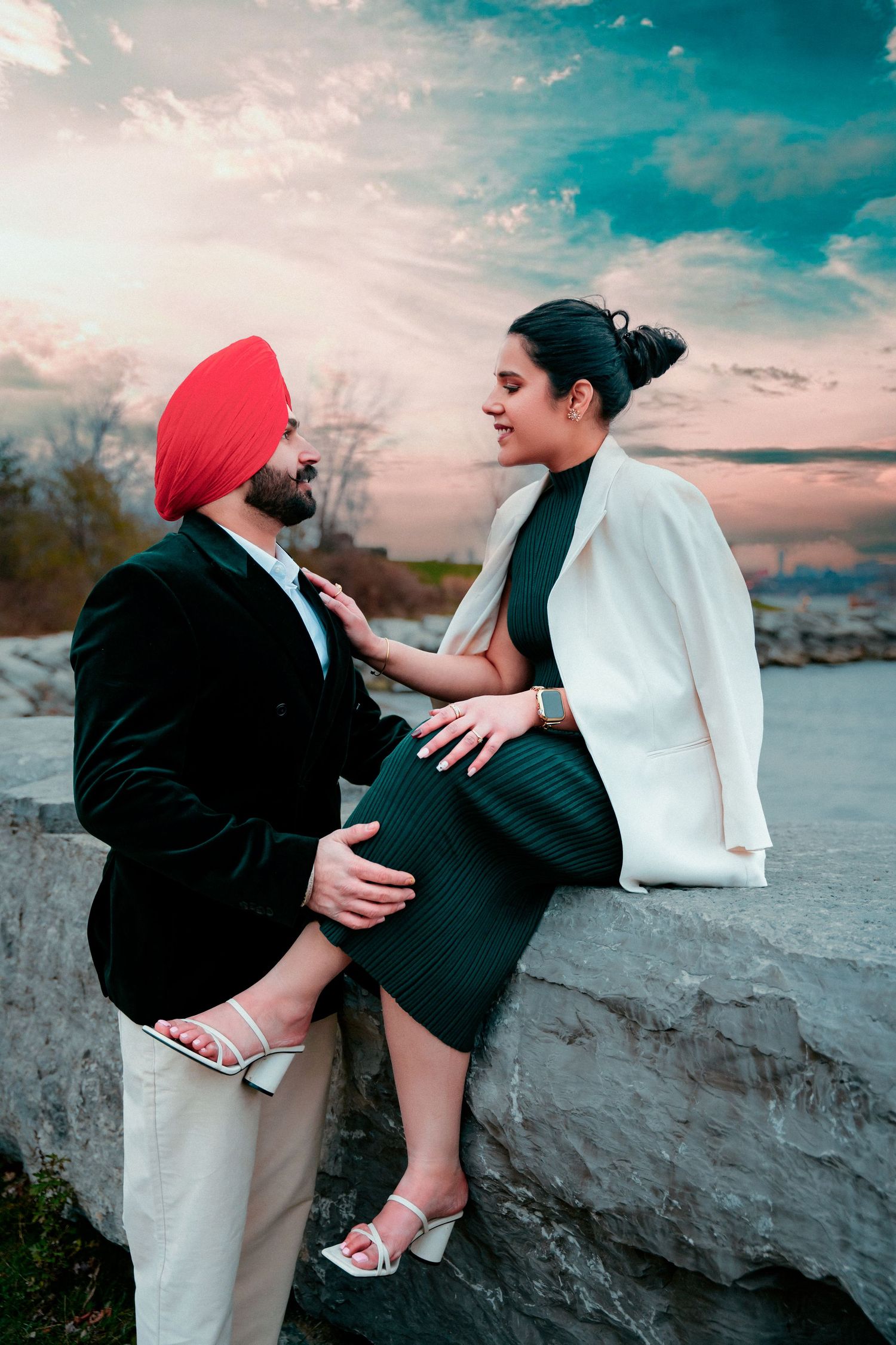 Couple shares intimate moments on rocky shoreline during sunset with urban backdrop.