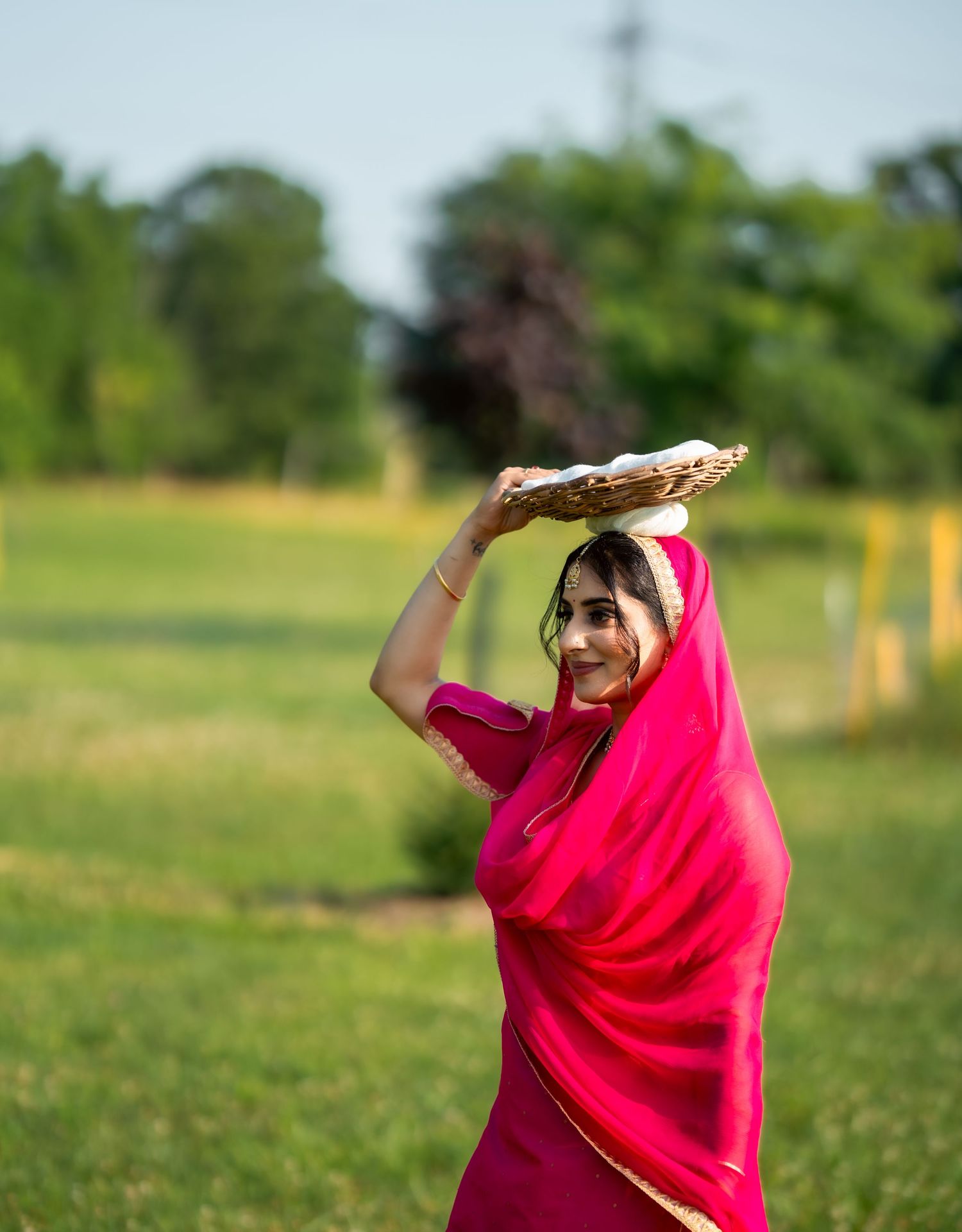A person in a vibrant pink traditional sari holds a decorative plate overhead while standing in a grassy field.