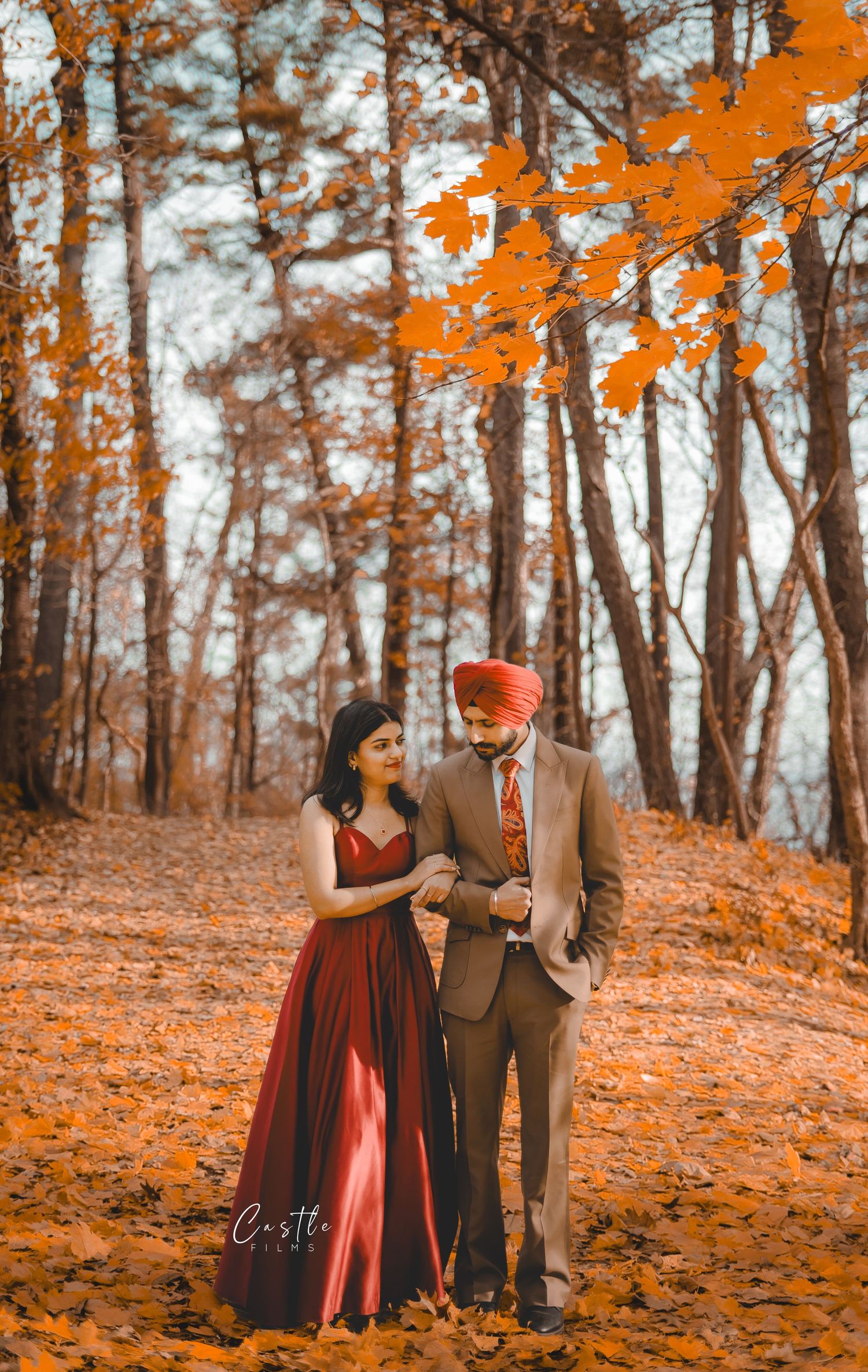 Couple in autumn forest with red dress and tan suit surrounded by vibrant orange fall foliage and leaves.