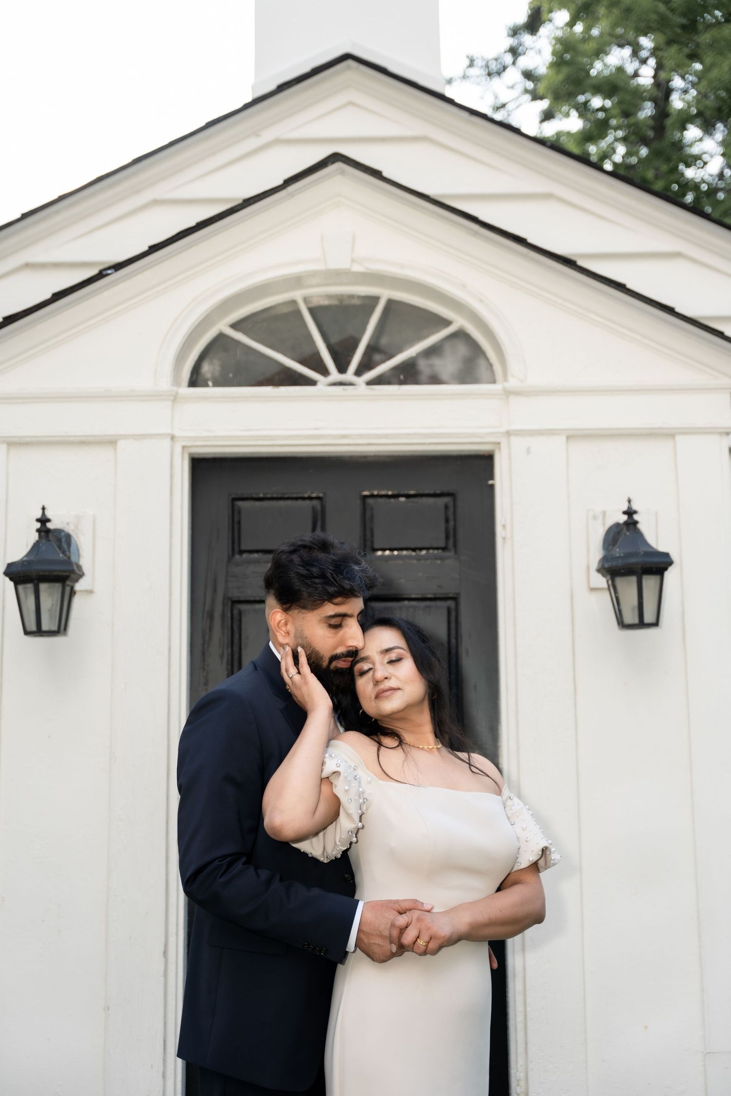 Romantic couple embraces in front of a white chapel with black doors and lanterns.