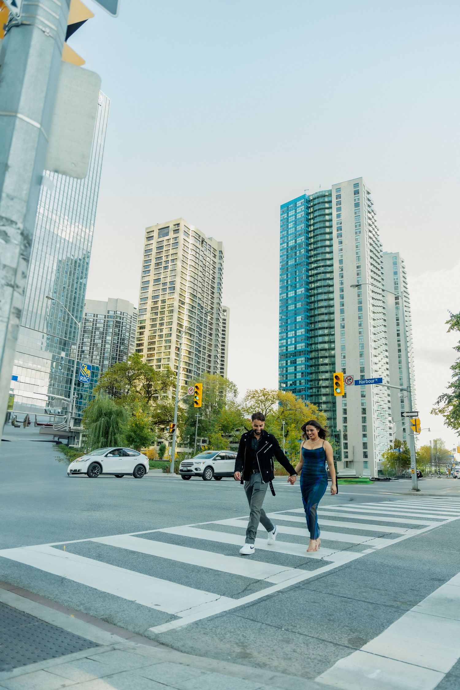 Two people cross a crosswalk in an urban setting with modern high-rise buildings and clear blue skies in the background.