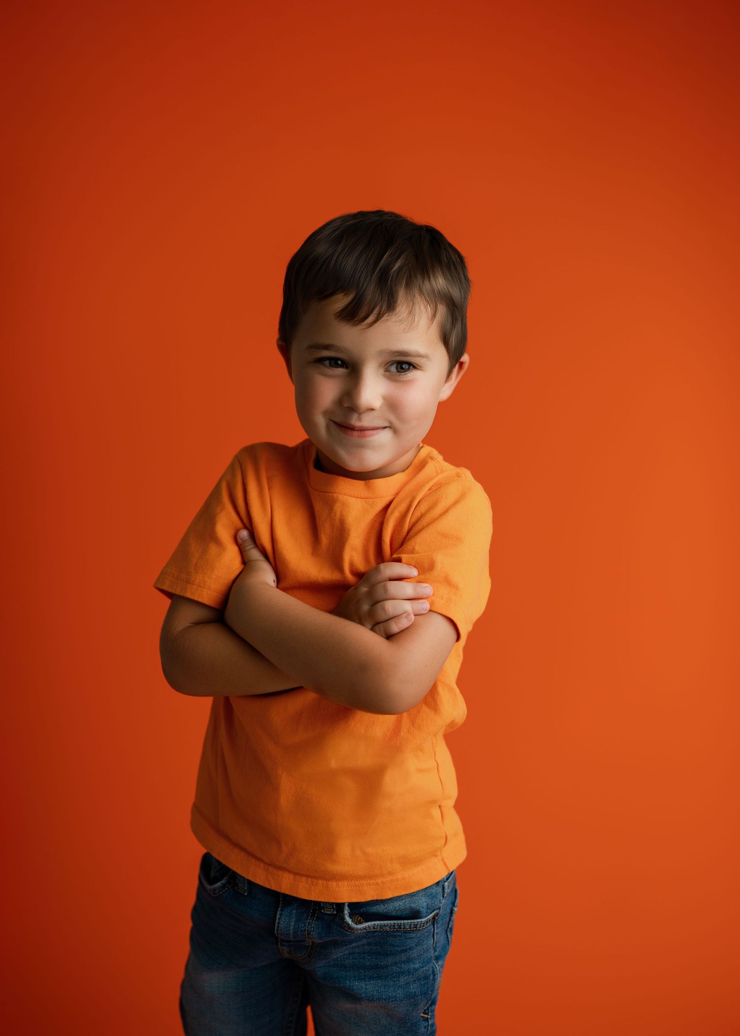 Young child wearing orange t-shirt and jeans poses against vibrant orange background in studio portrait series.