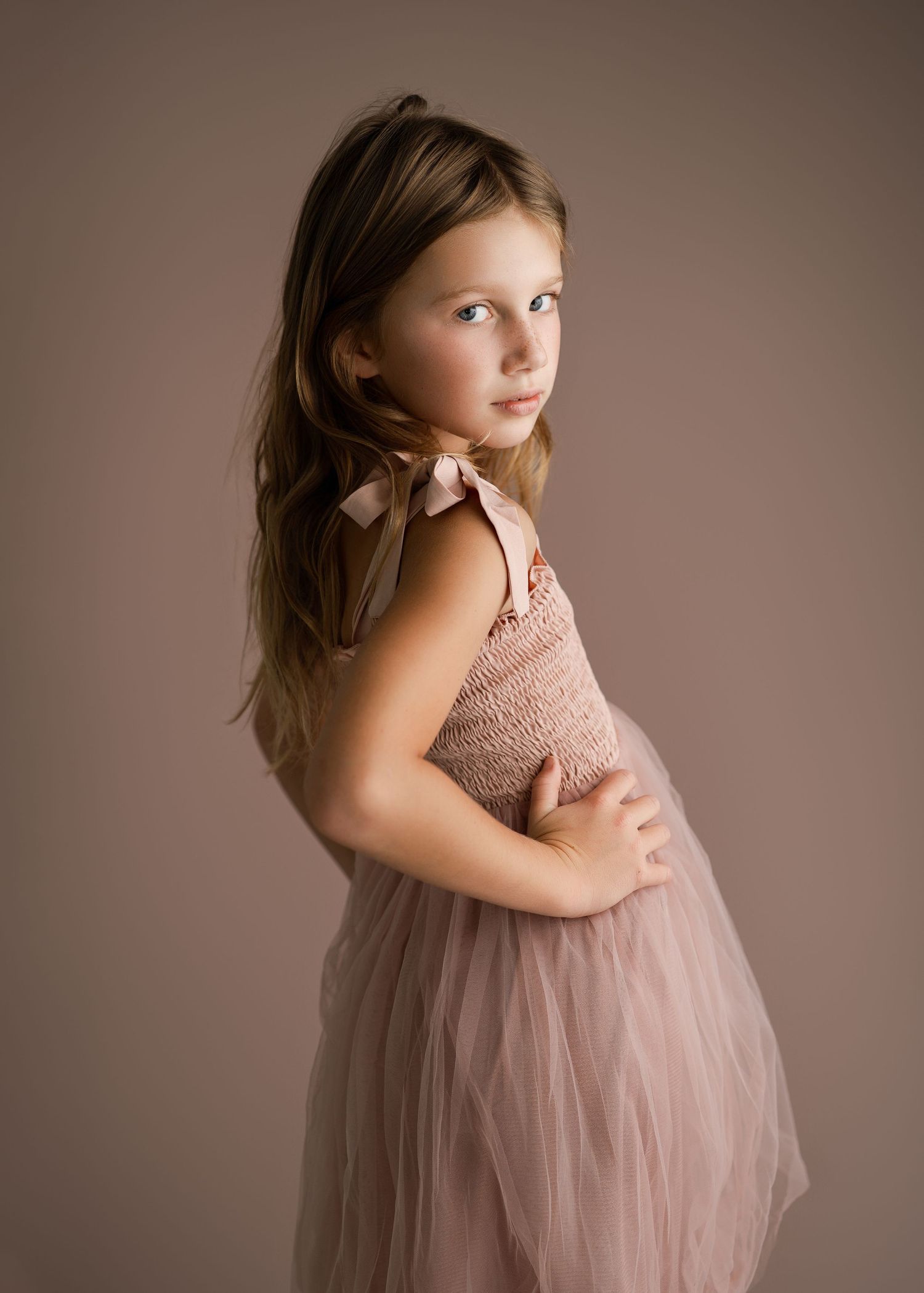 Young dancer in blush pink tulle dress poses gracefully against neutral studio background.