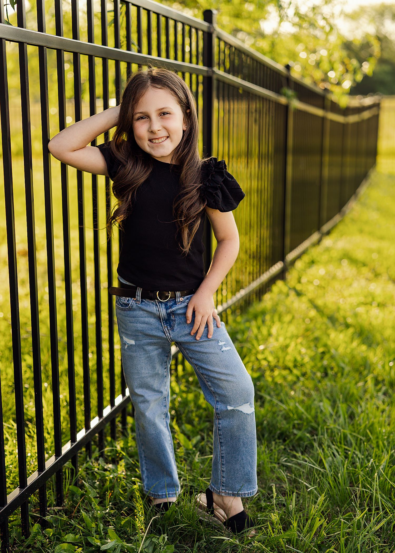 Child in black shirt and jeans poses playfully by metal railing in sunlit grassy area.