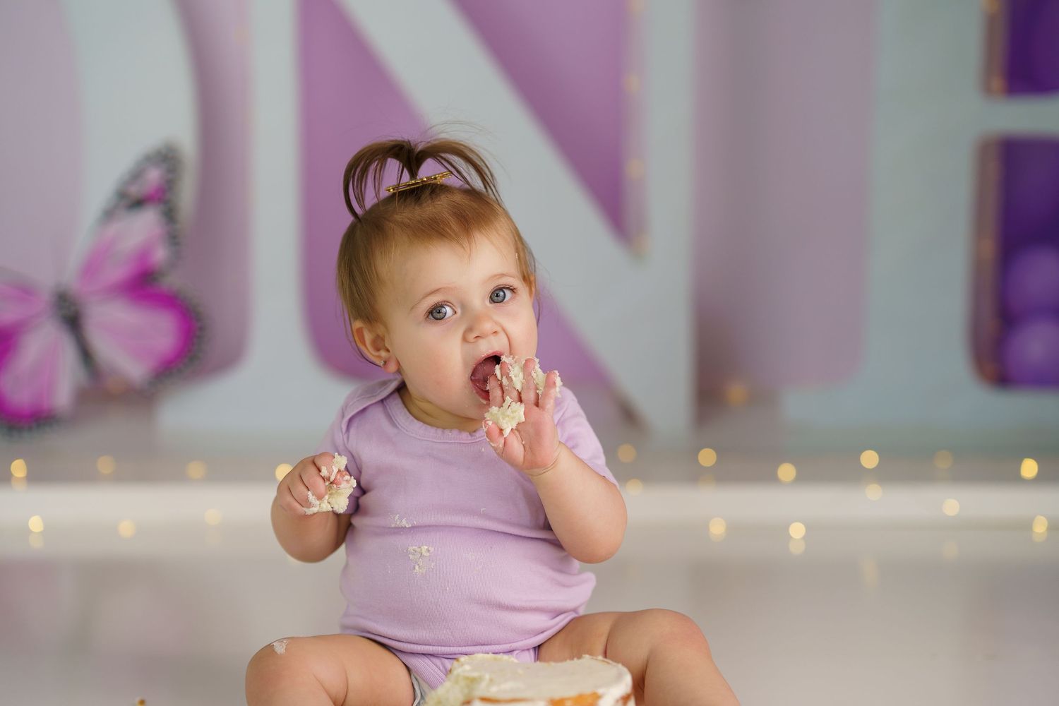 Baby in purple outfit enjoying cake smash photo session with butterfly decorations in background.