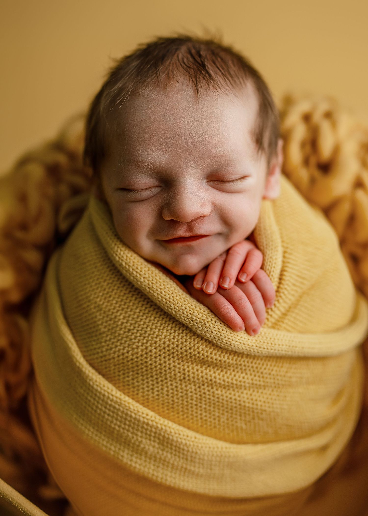 Sleeping newborn wrapped in soft yellow knit blanket against warm golden backdrop.