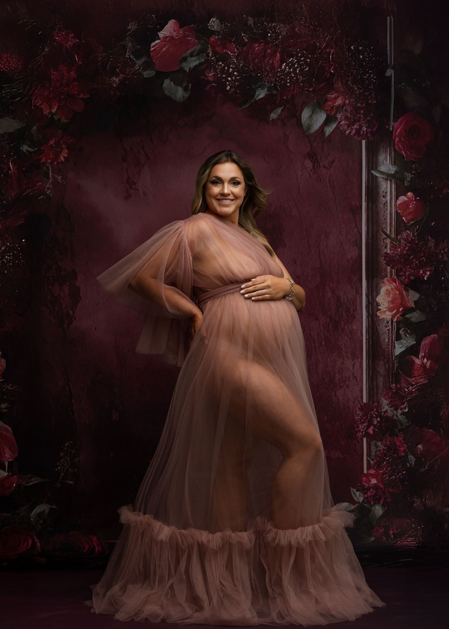 Dreamy maternity photoshoot with flowing pink tulle dress against floral backdrop.