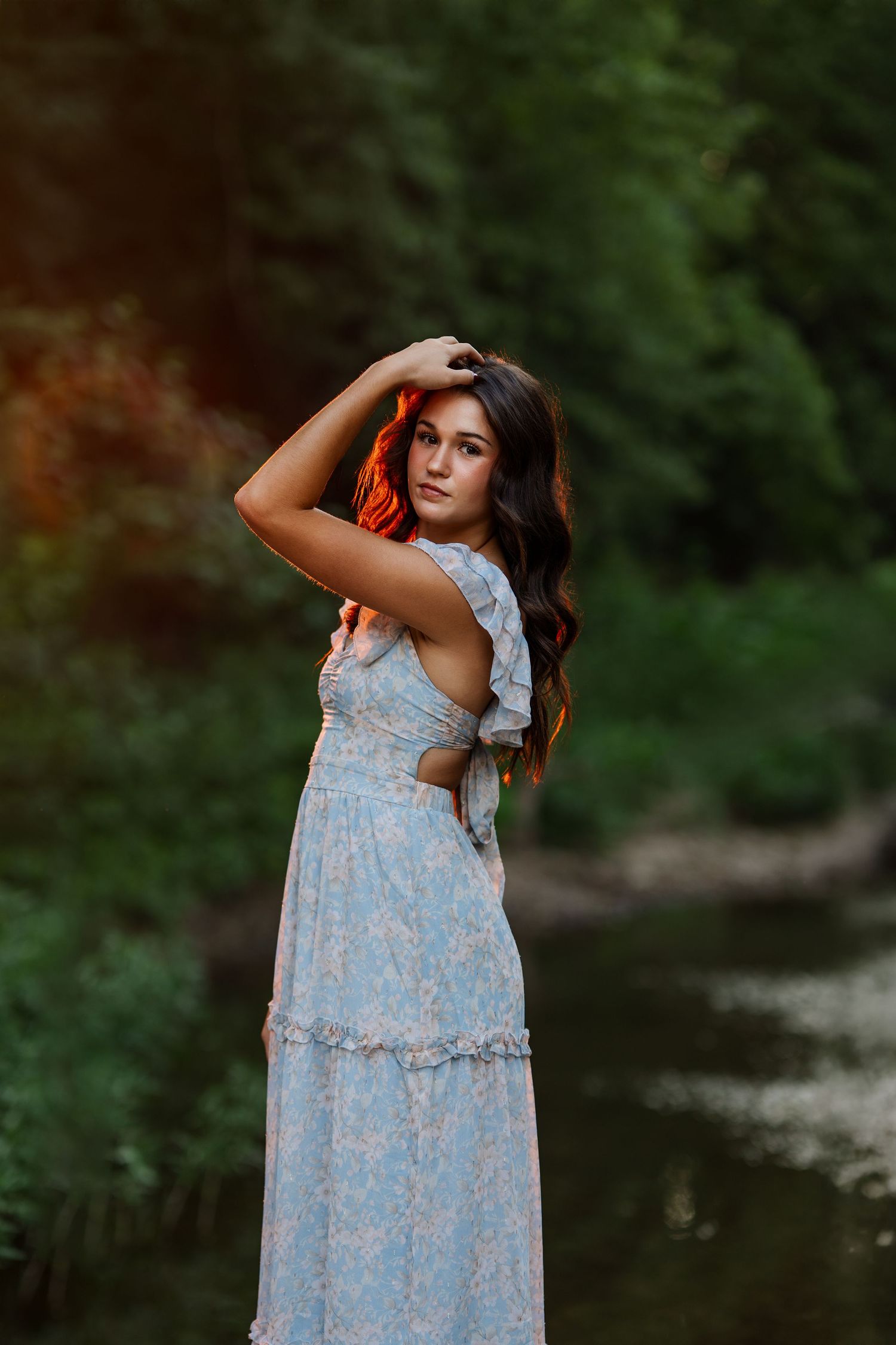 Person in light blue floral dress poses by water in natural outdoor setting.