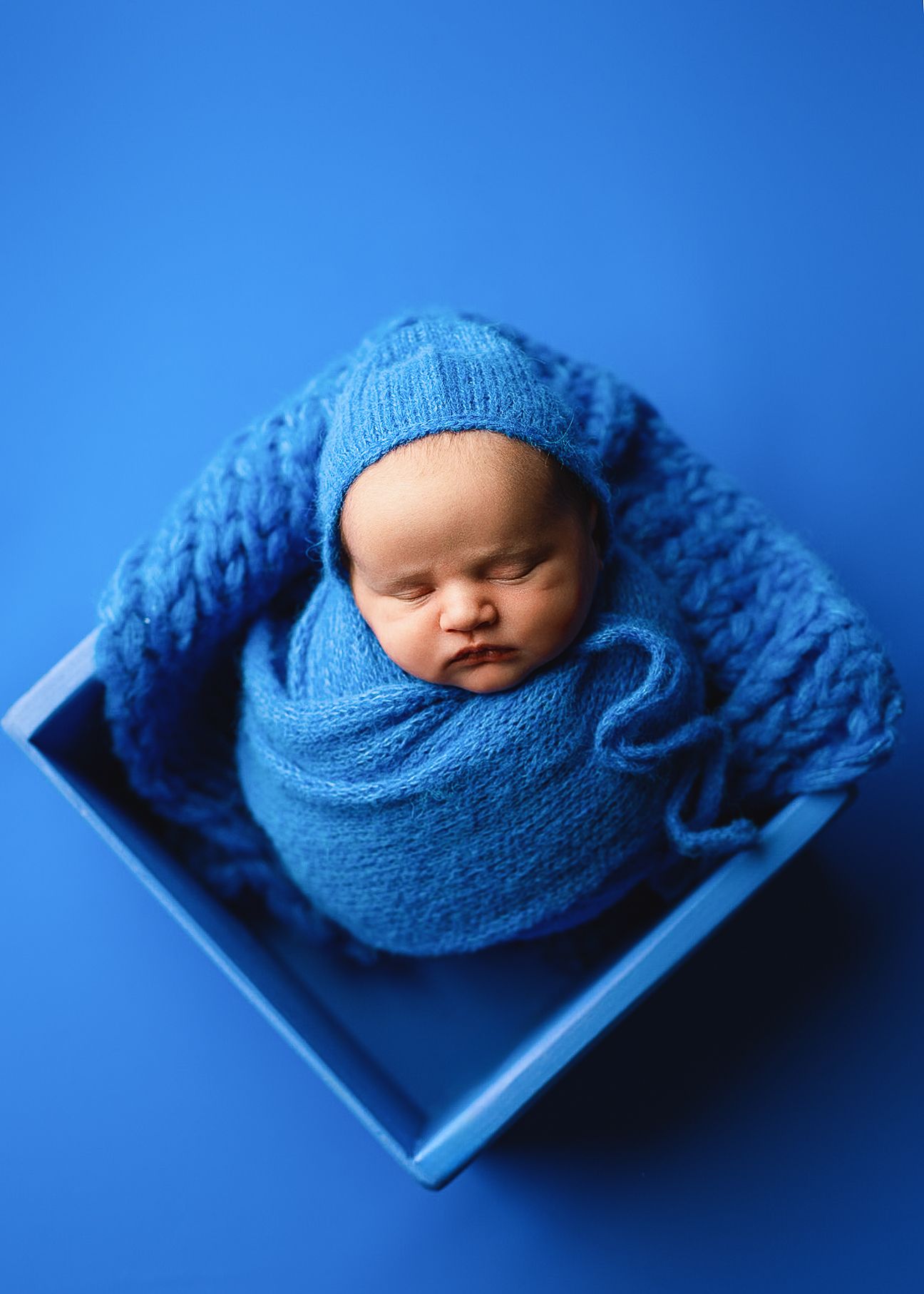 Newborn baby wrapped in blue knit blanket sleeps peacefully against blue backdrop.