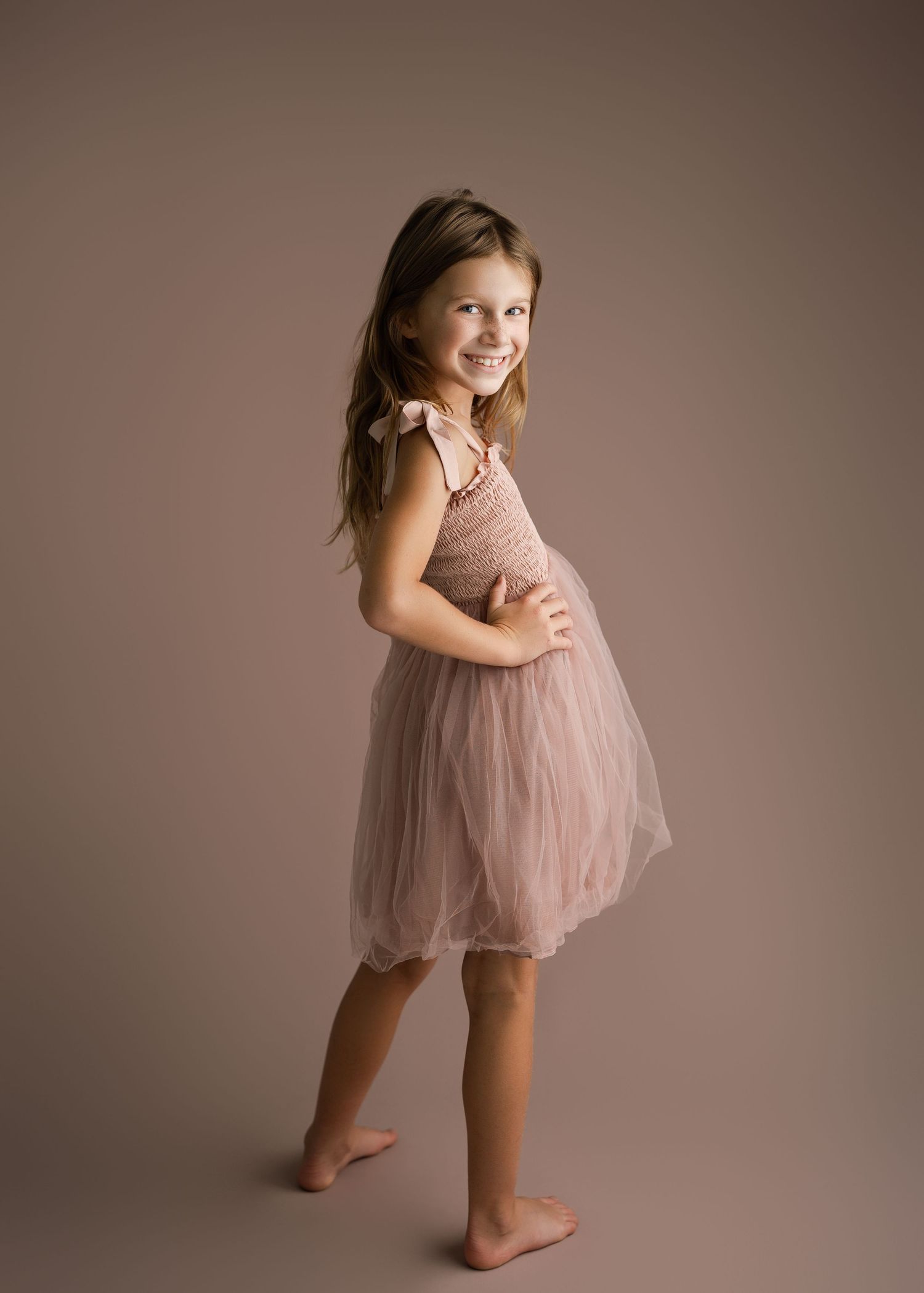 Child performs dance movements in pink tulle dress against neutral studio background.