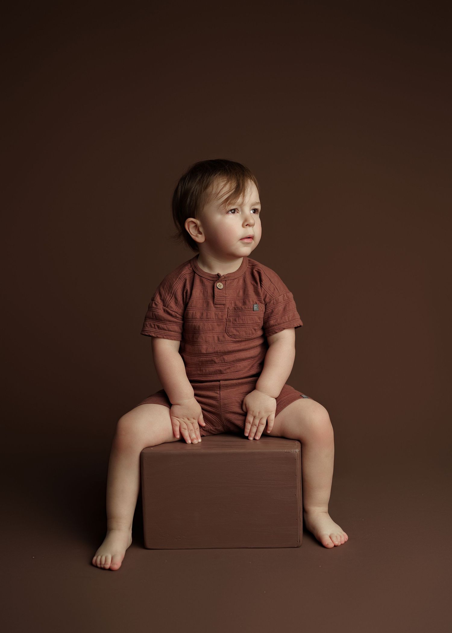 Baby in brown shirt sitting on dark platform during professional photo session.