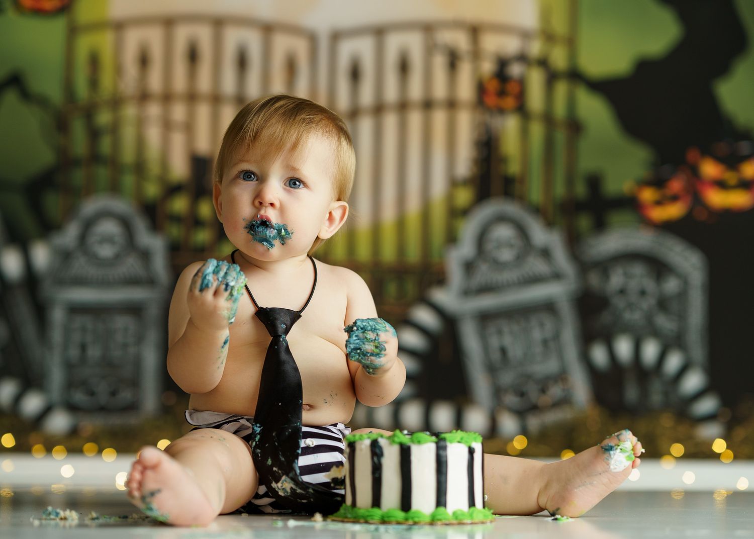 Festive Halloween-themed cake smash photo session with black and white striped outfit against spooky graveyard backdrop.