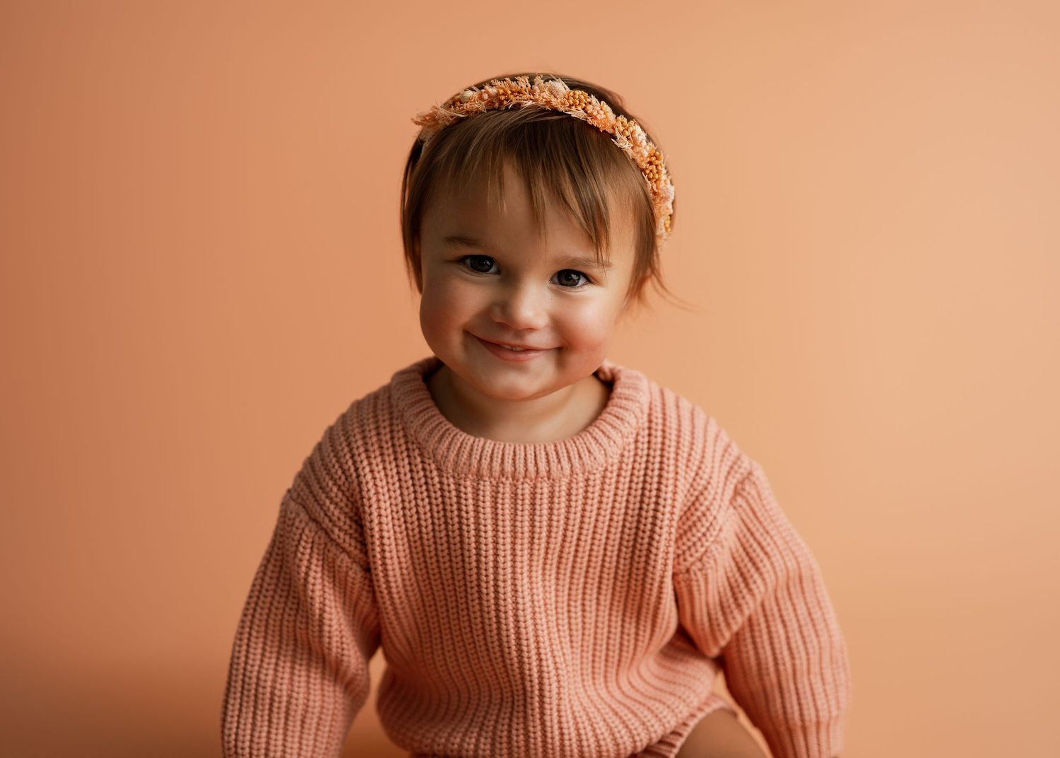 Child wearing peach-colored sweater poses against warm orange background.