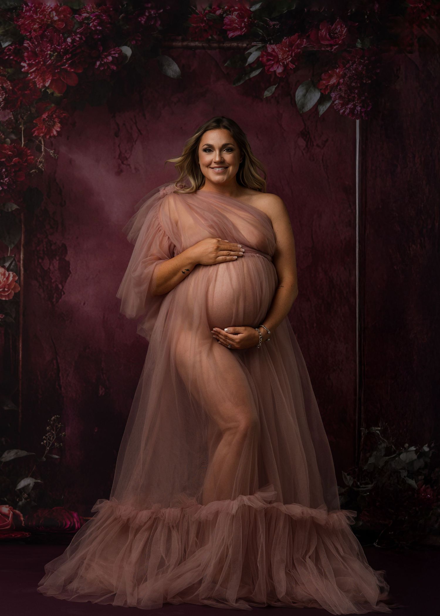 Romantic maternity portrait with pink tulle gown surrounded by floral arrangements.