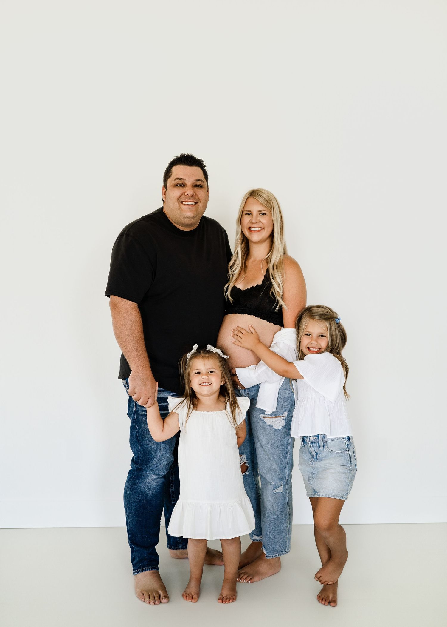 Family portrait with expecting mother in casual attire against white studio backdrop.