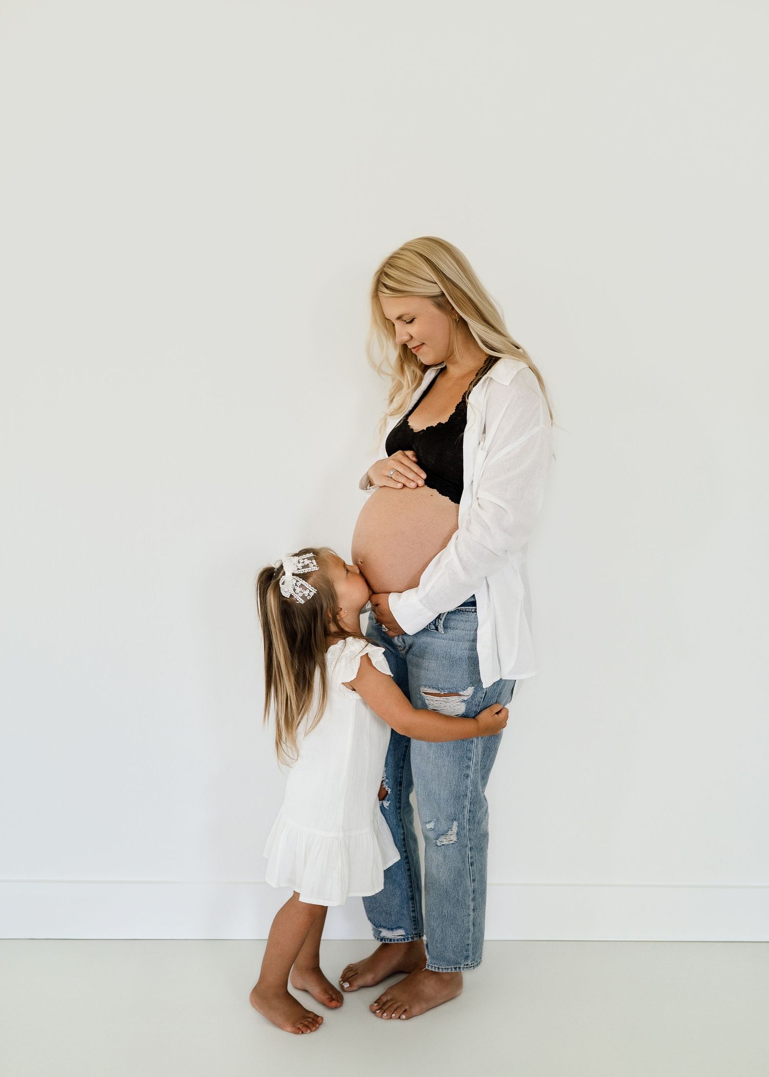 Maternity photo of pregnant woman in white shirt and jeans embracing young child against white wall.