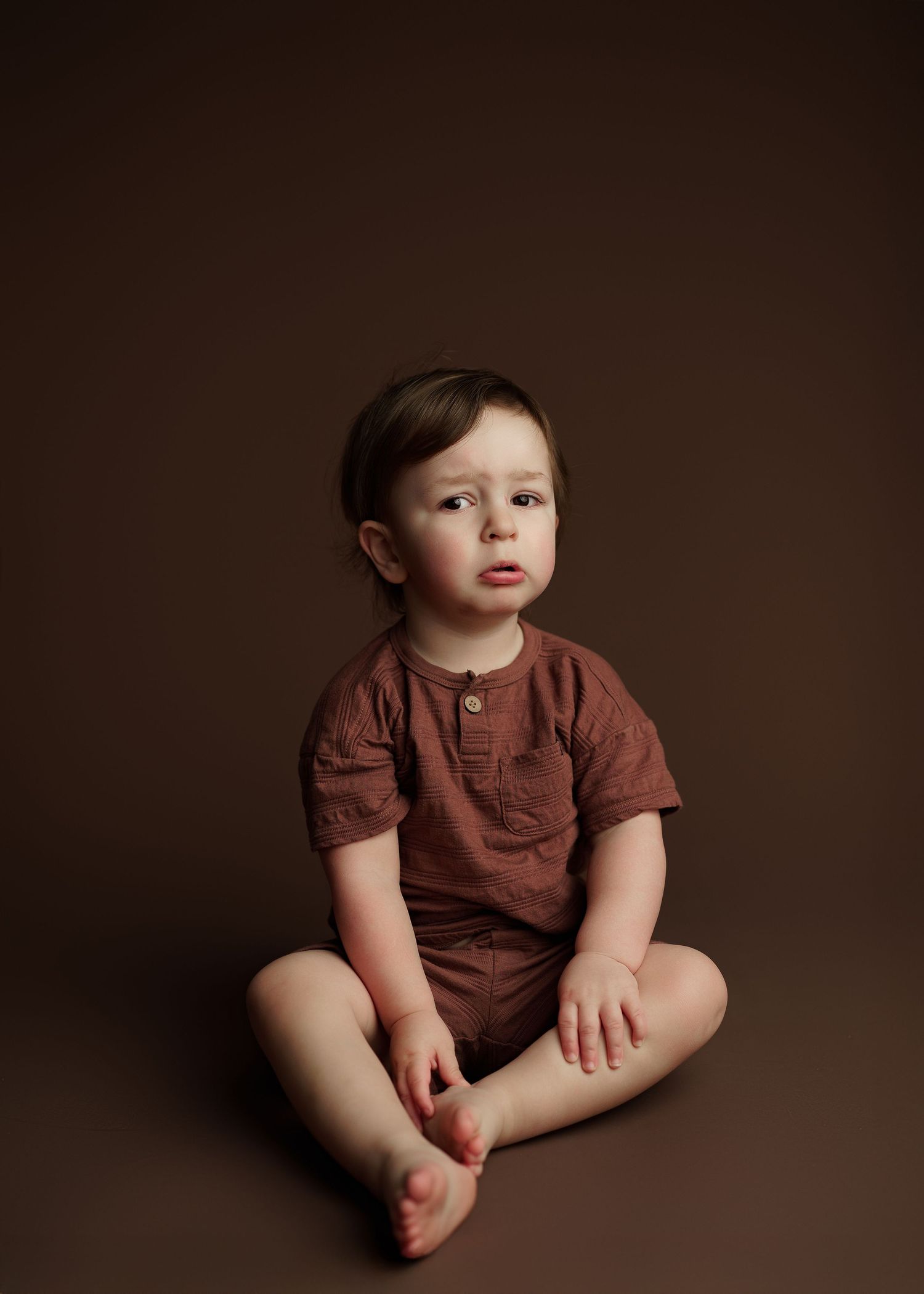 Baby wearing brown t-shirt sitting against dark background in studio setting.