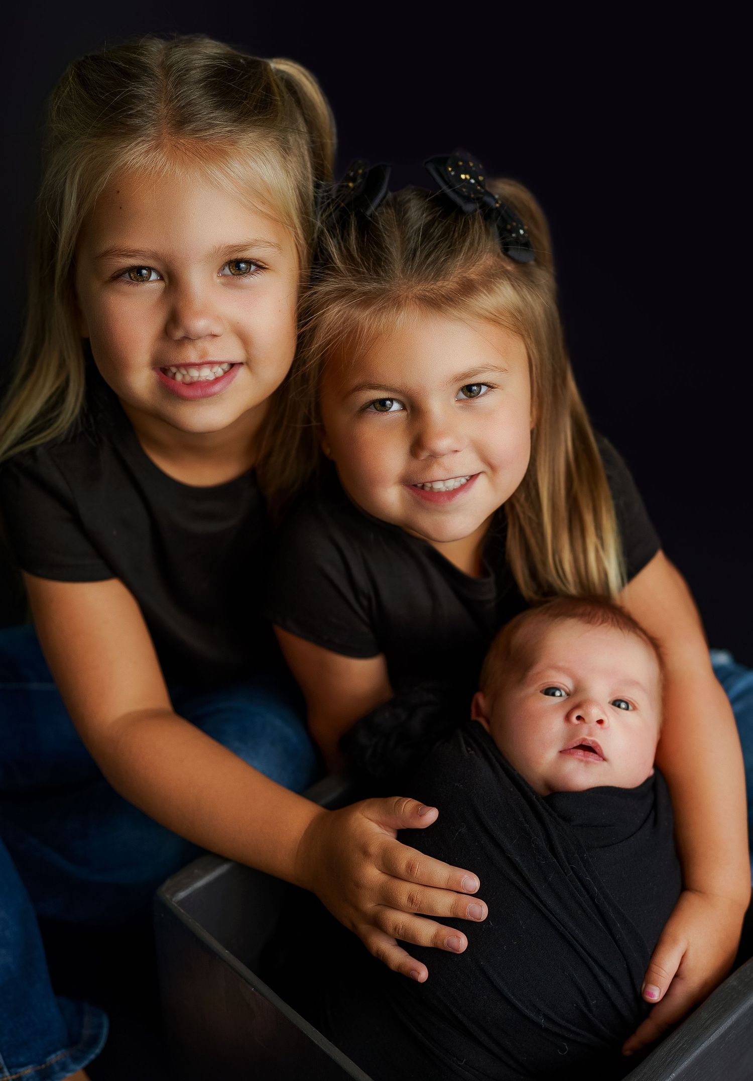 Family portrait against dark background showing siblings bonding with new baby.