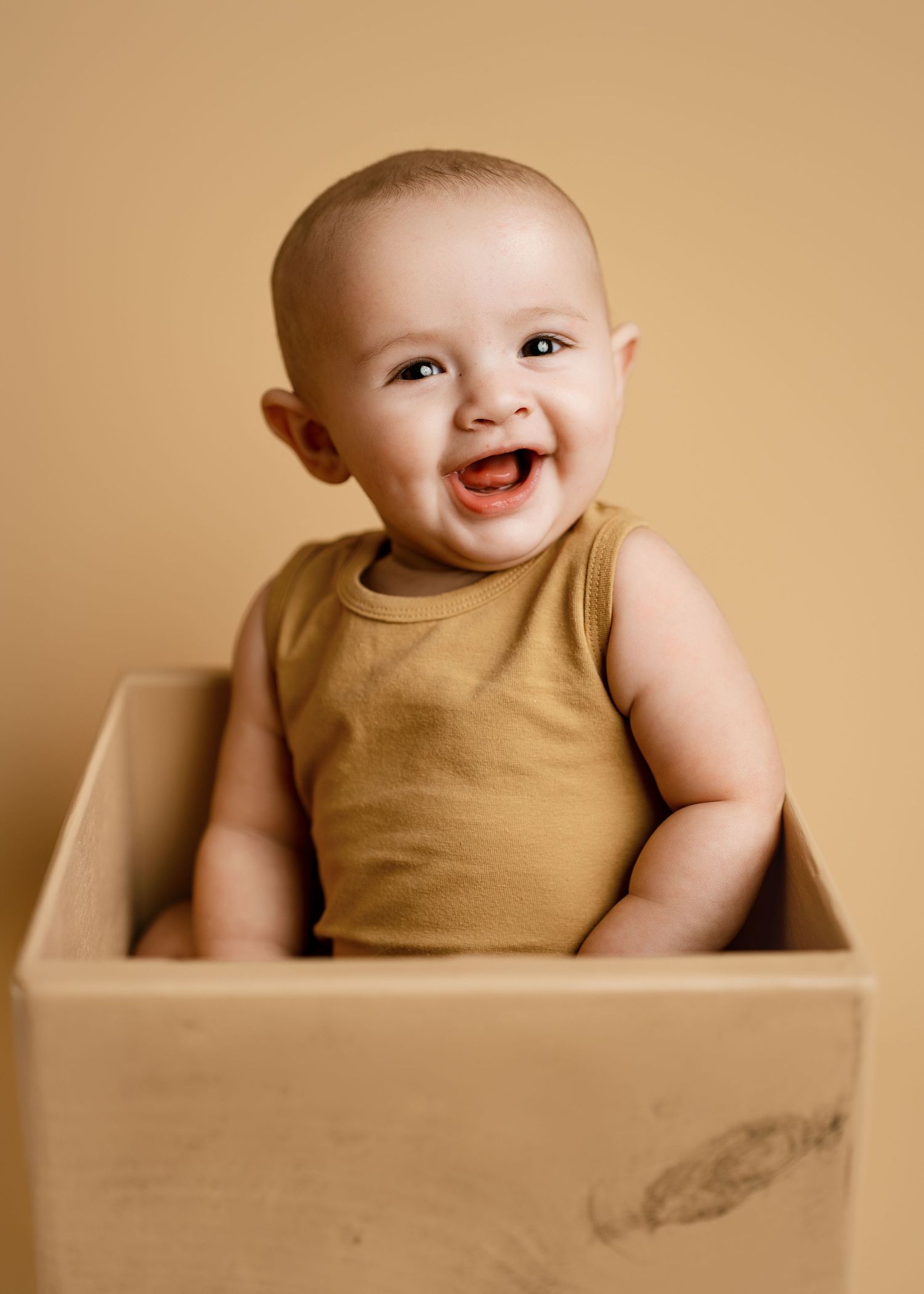 Happy baby sitting in wooden box wearing tan outfit against beige background.