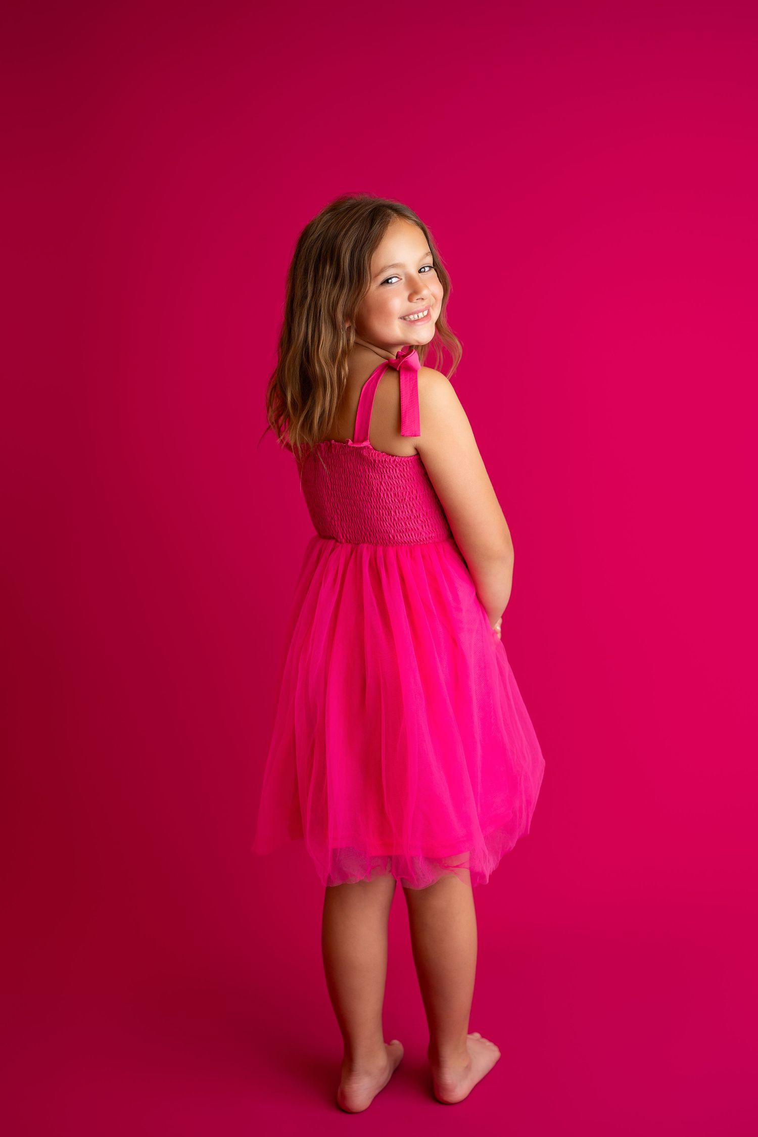 Dancer in flowing pink dress kneels gracefully on floor against fuchsia backdrop.