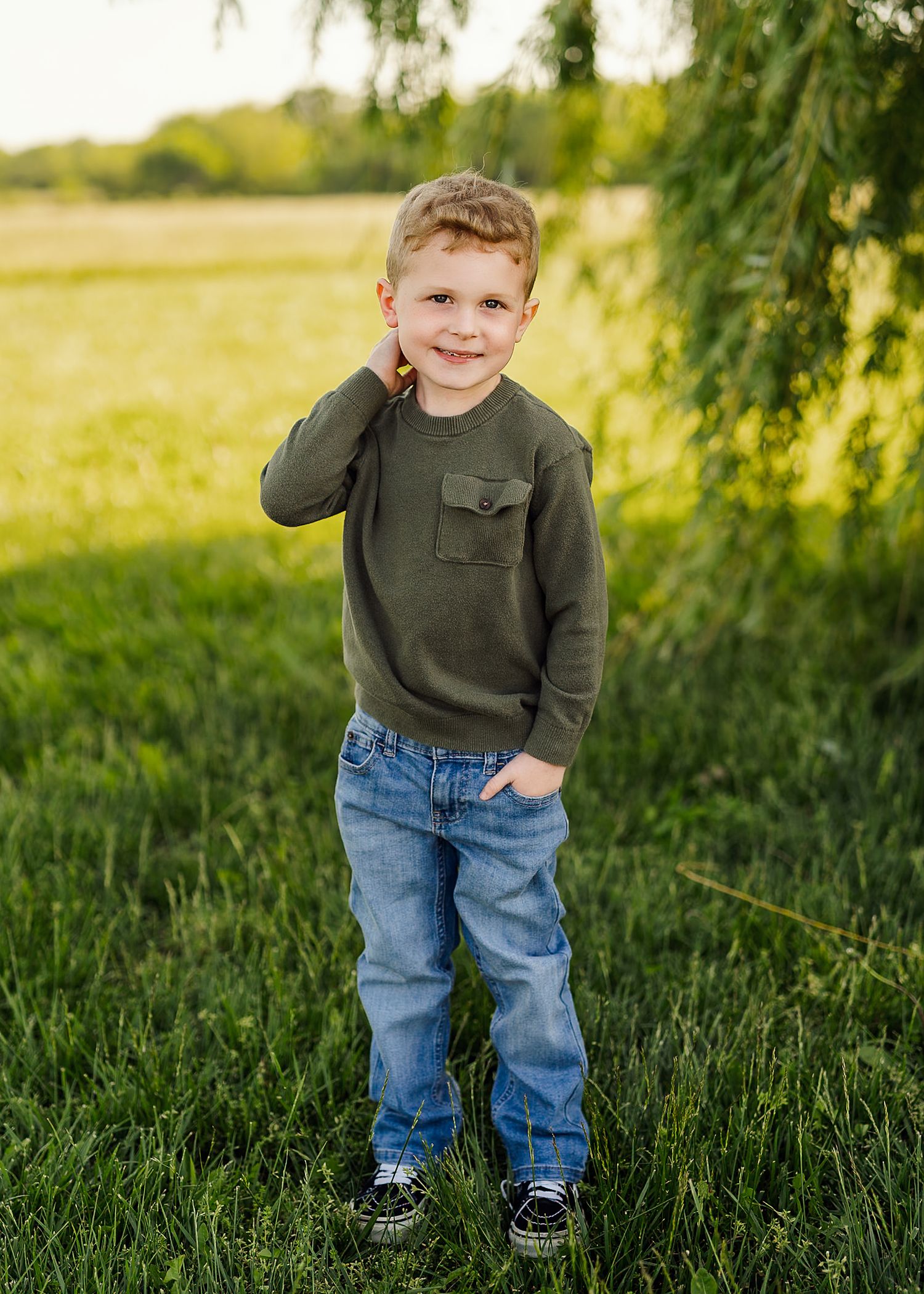 Child in green sweater and jeans posing playfully in sunny garden setting.