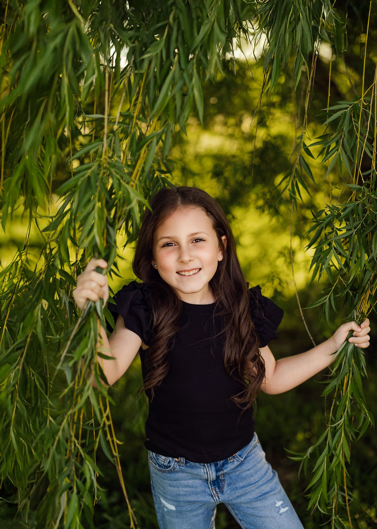 Young child playing under drooping willow tree branches in a black shirt and jeans on a sunny day.
