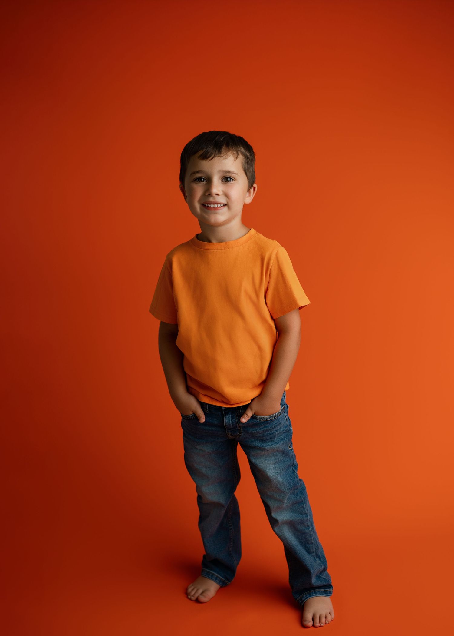 Child in orange t-shirt and jeans poses playfully against bright orange background.