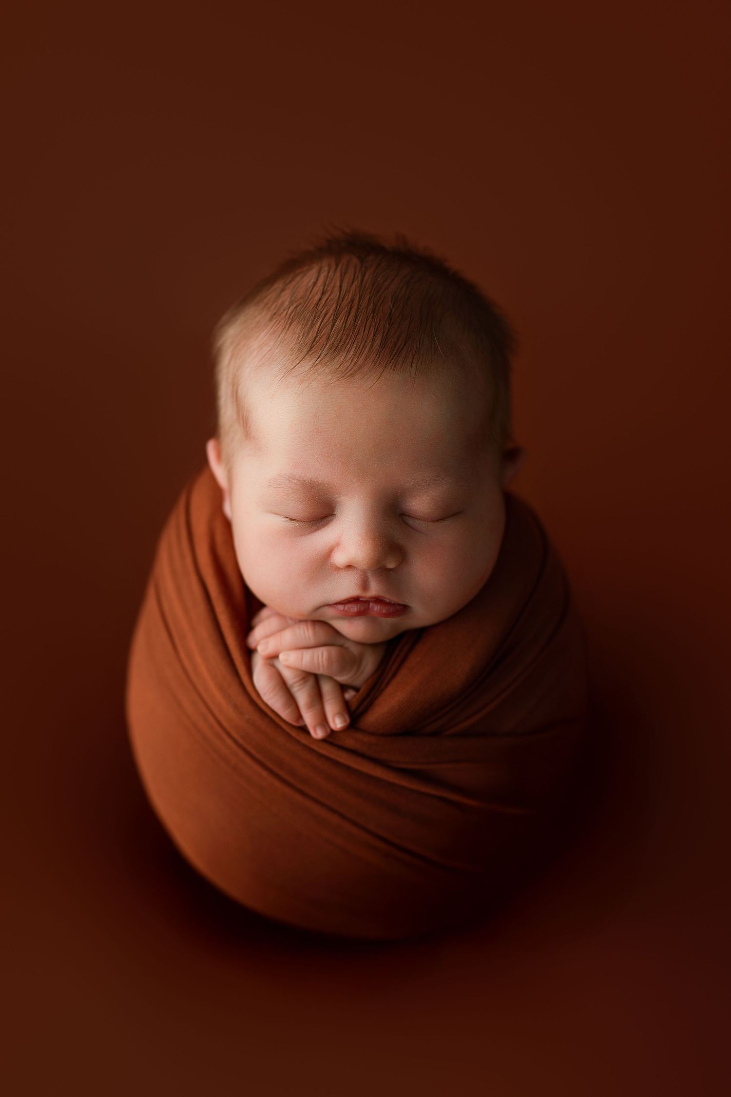 Sleeping newborn wrapped in brown fabric against brown background.