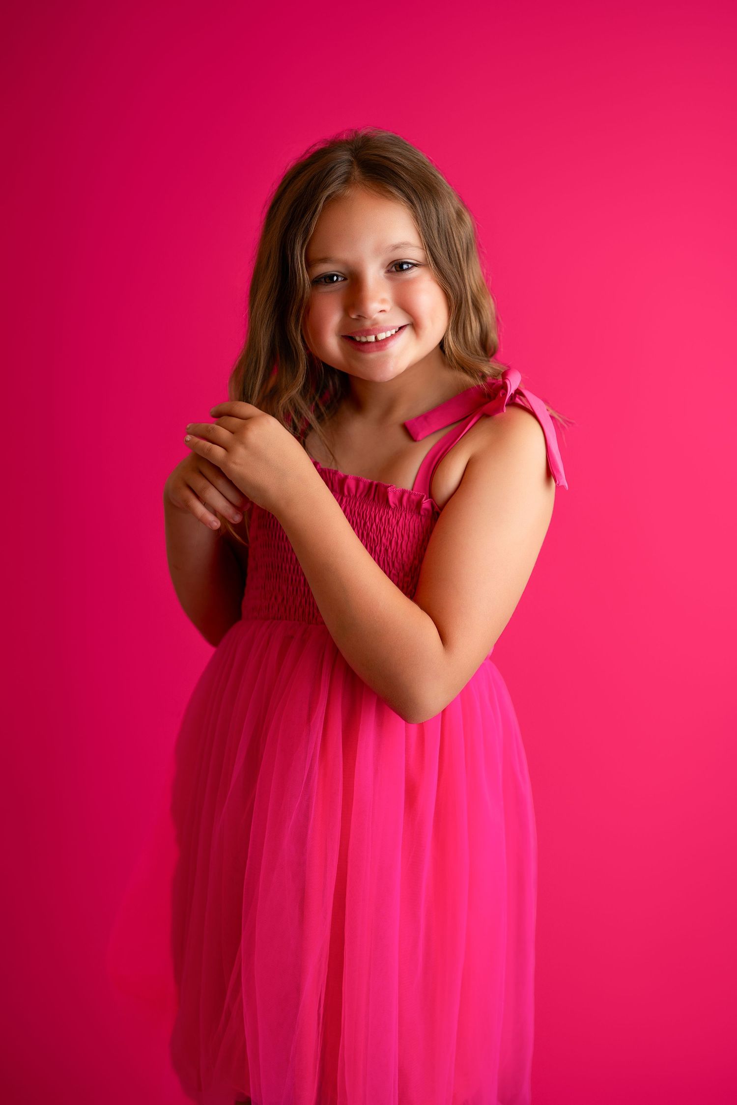 Young dancer in bright pink tulle dress poses playfully against vibrant pink backdrop.