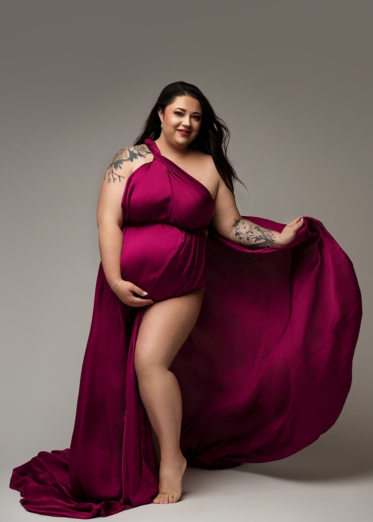 One-shoulder burgundy gown with flowing fabric creates elegant silhouette against grey backdrop.