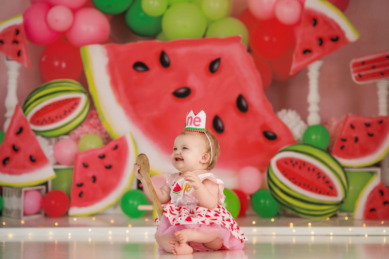 A watermelon-themed backdrop with pink and green decorations and string lights for a party setting.