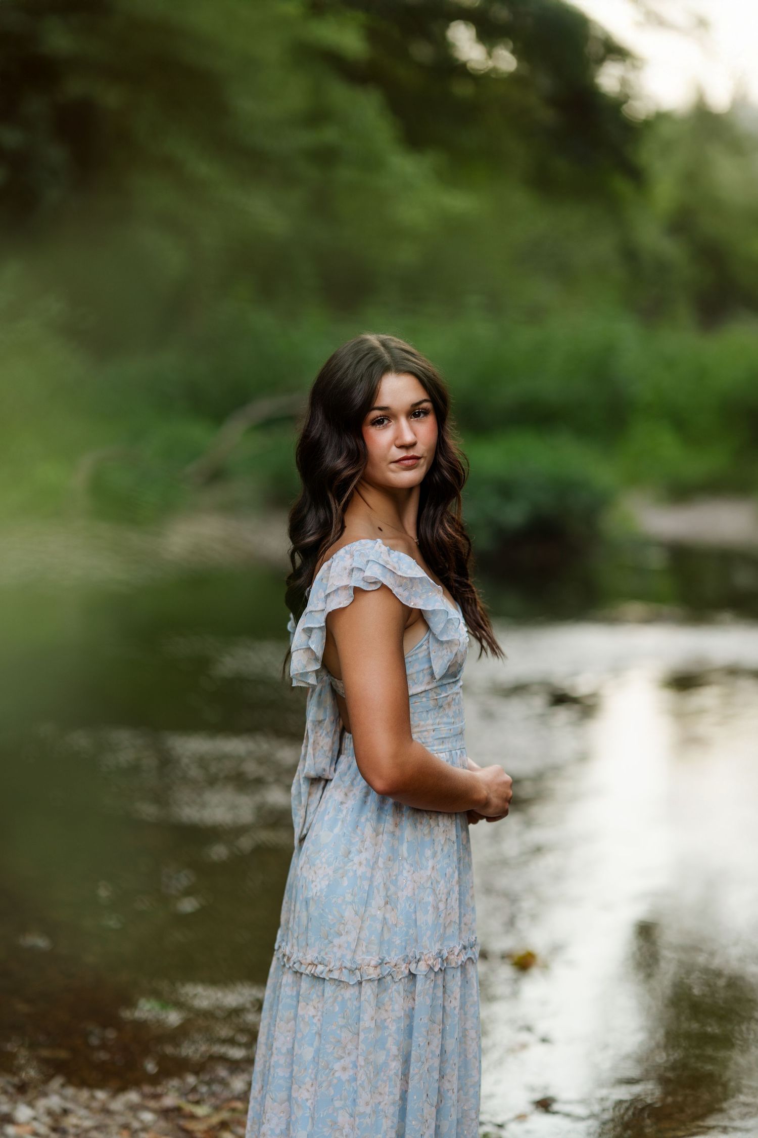 Subject wearing light blue summer dress stands near peaceful waterside.