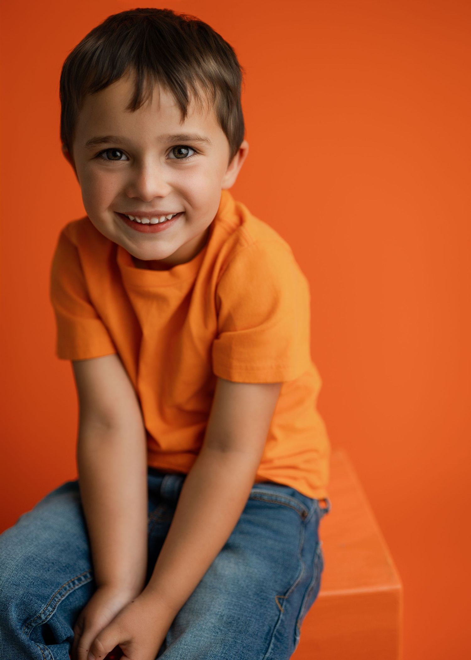 Child wearing orange t-shirt and blue jeans poses playfully on orange studio background.