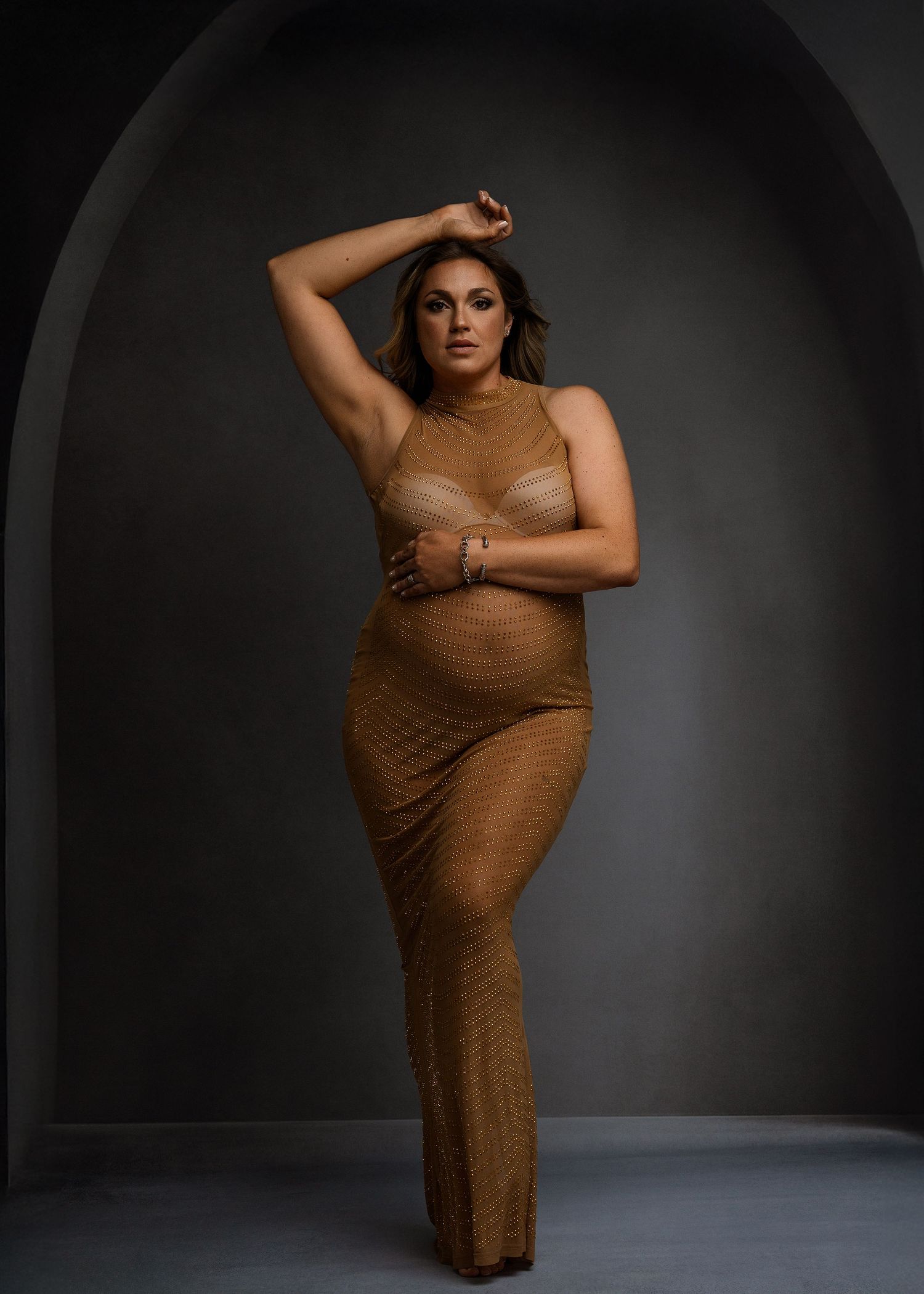 Maternity photoshoot in brown fitted dress against dark dramatic background.
