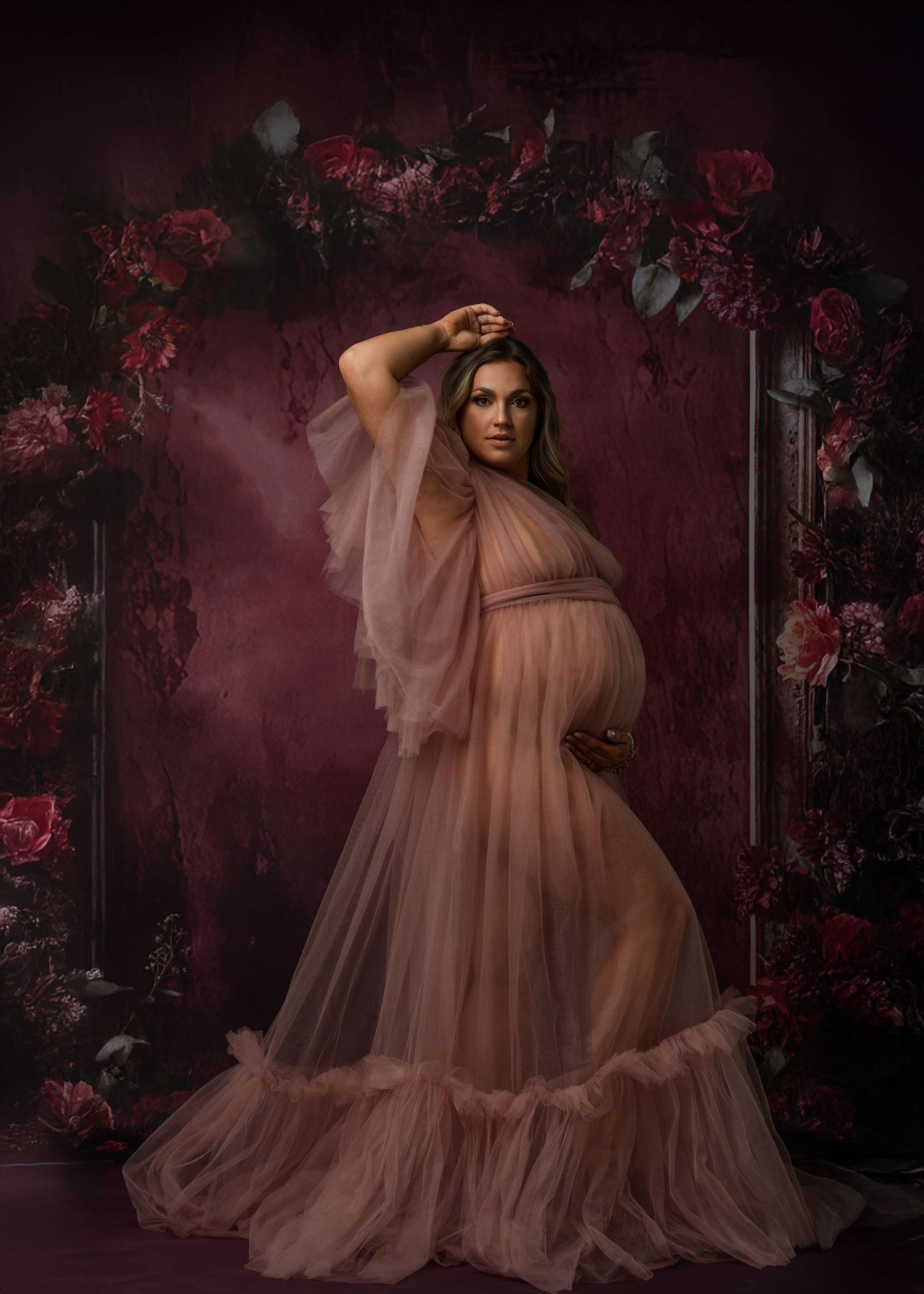 Dreamy maternity photos with pink tulle dress against dark floral backdrop.