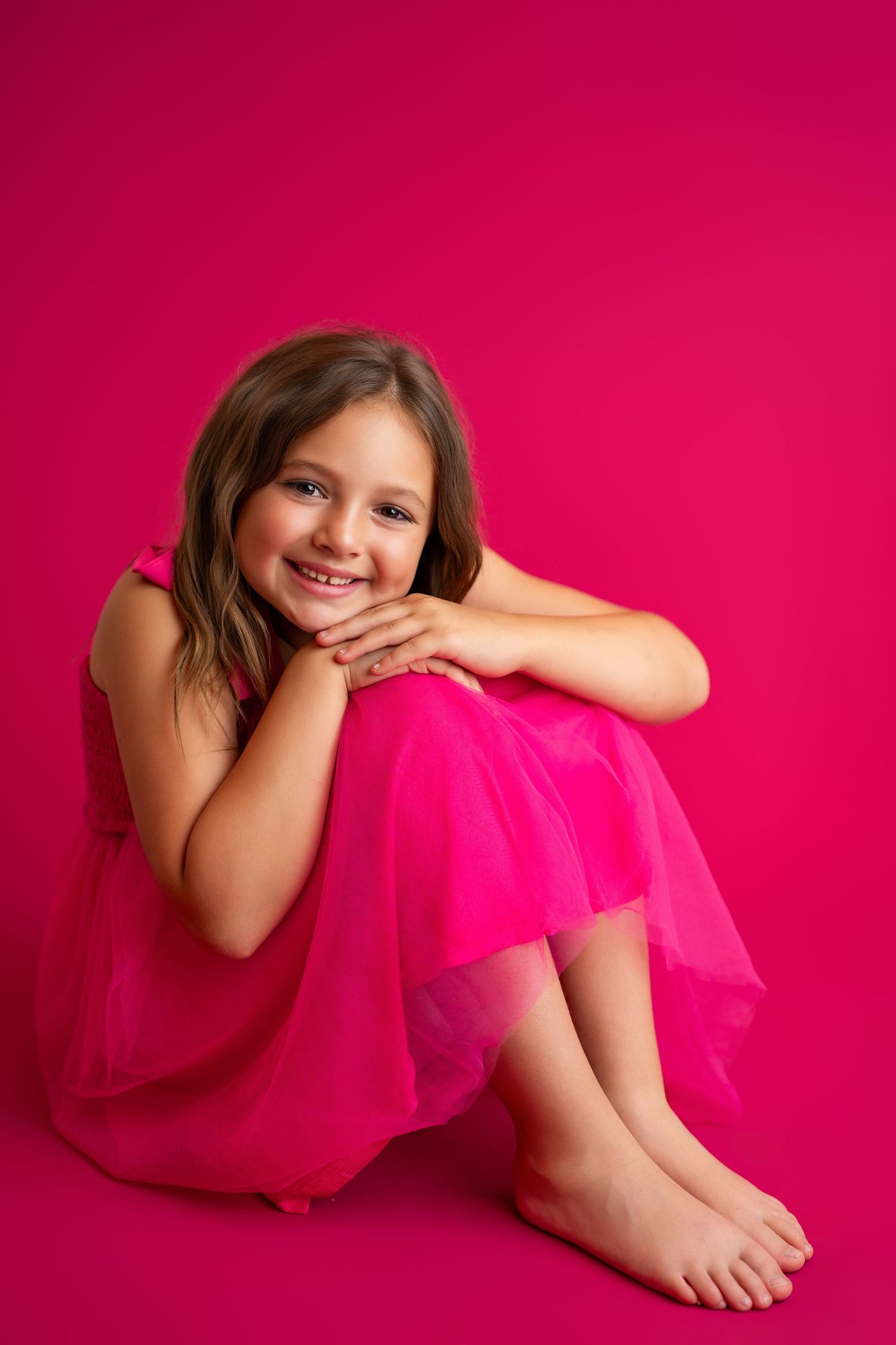Young girl in pink tulle dress sitting against bright pink backdrop.
