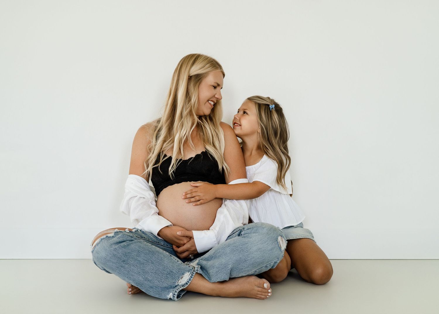 Mother and daughter sharing intimate moment during maternity photoshoot against white wall.