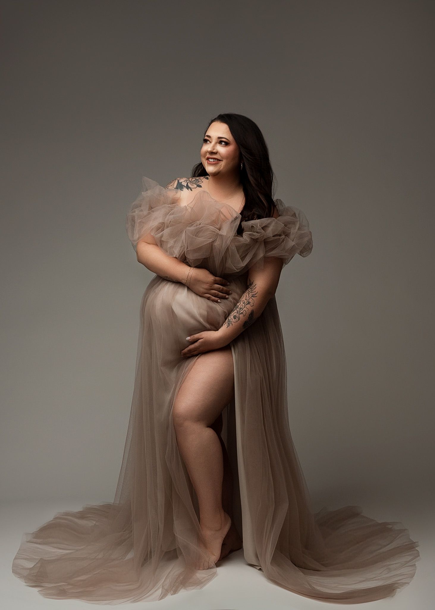 Ethereal beige tulle maternity gown with ruffled shoulders flows gracefully in studio portrait.
